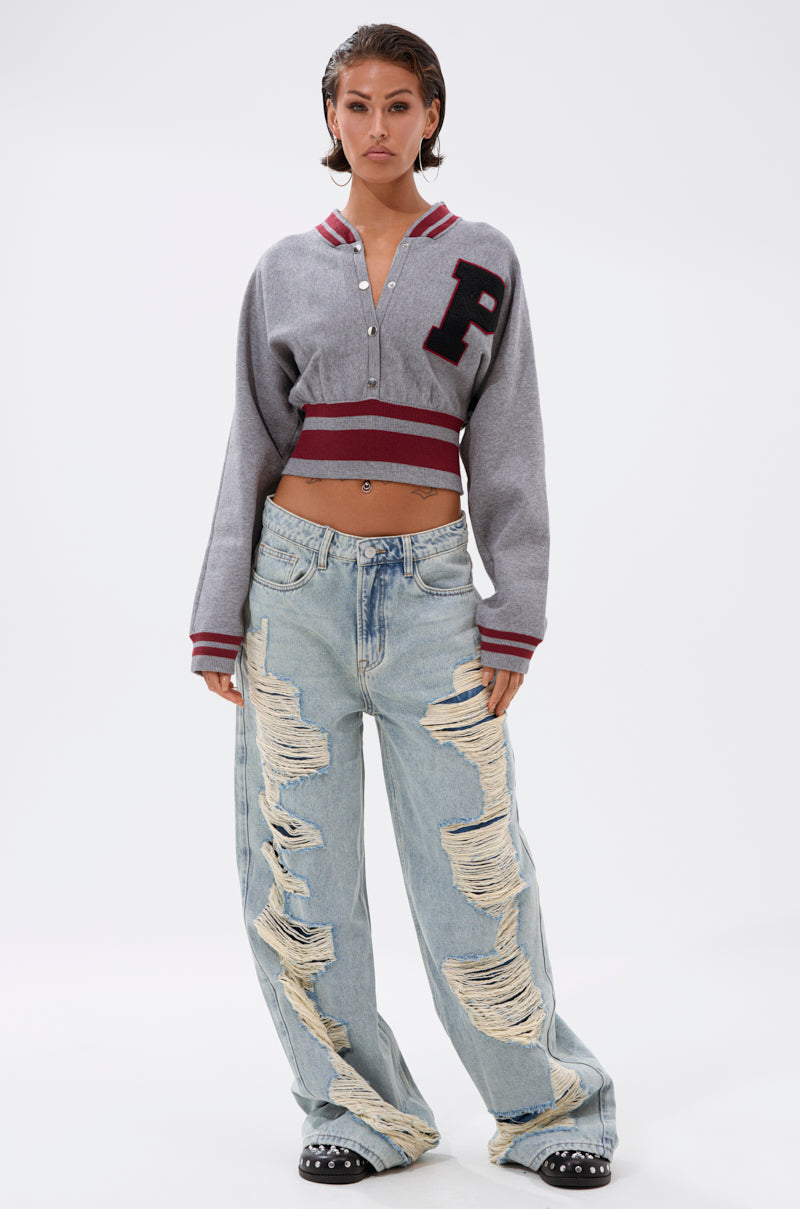 Facing forward against a white background, a person with short hair wears the Varsity Team Button Down Sweater in Heather Grey, featuring a bold "P" patch, paired with light blue distressed wide-leg jeans. 6