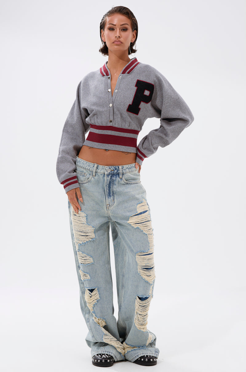 A woman stands confidently in the Varsity Team Button Down Sweater in Heather Grey, paired with baggy light-wash distressed jeans and studded black shoes, set against a plain white background. 7