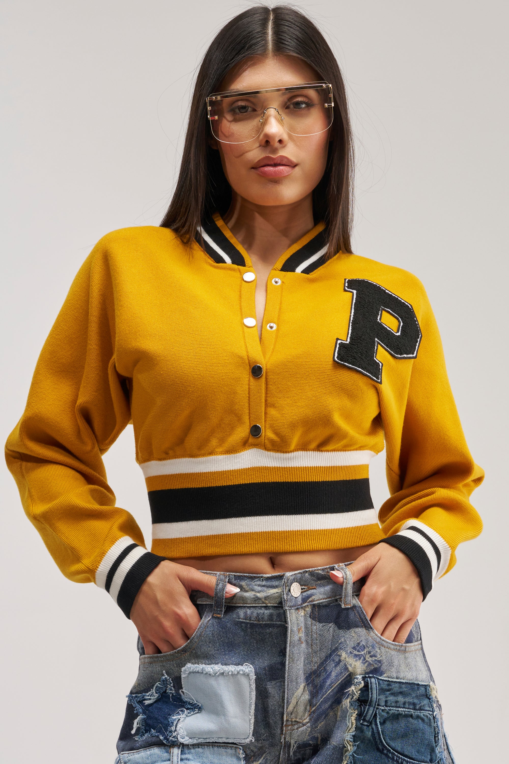 Wearing the VARSITY TEAM BUTTON DOWN SWEATER IN MUSTARD—a yellow varsity-style cropped cardigan with a "P" patch, striped cuffs and hem—a person stands with hands in pockets against a light background, paired with clear glasses and distressed denim jeans.