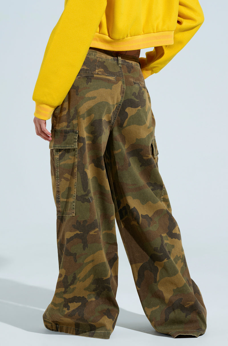 A person wearing a yellow sweatshirt and HIDDEN IN PLAIN SIGHT CARGO PANT stands with their back to the camera. These camo print, high-rise cargo pants feature multiple pockets and are set against a light blue background.