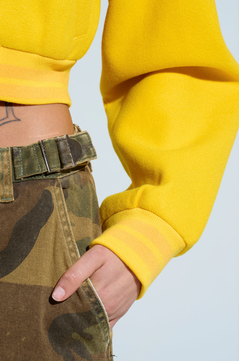 A person wearing a bright yellow long-sleeve top with their right hand in the pocket of their high-rise "Hidden In Plain Sight" cargo pants, featuring a camo print. A small tattoo is visible on their torso above the waistband.