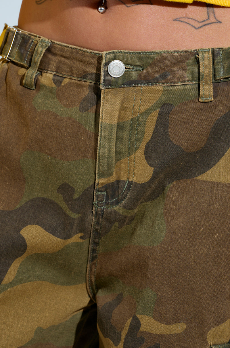 A close-up shot features someone wearing the HIDDEN IN PLAIN SIGHT CARGO PANT, showcasing its high-rise camo print design with a visible button and belt loops. Adding intrigue to the look are hints of tan skin and a tattoo above the pants, set against a softly blurred blue background.