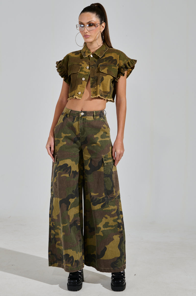 A person stands against a light background wearing a camouflage outfit with RIDE WITH ME CAMO WIDE LEG PANT featuring cargo pockets, complemented by a cropped jacket with ruffled sleeves, sunglasses, and black shoes.