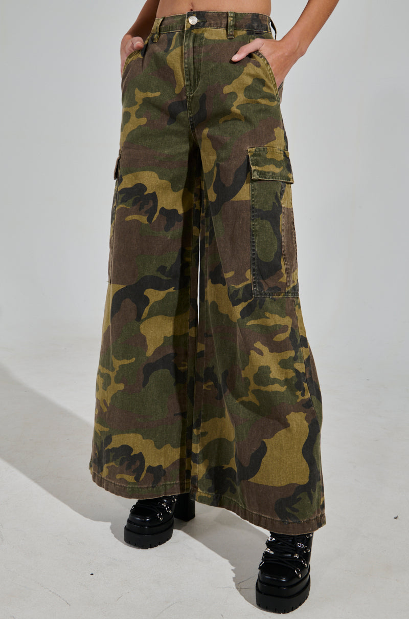 Someone wears the "RIDE WITH ME CAMO WIDE LEG PANT" featuring high-rise cargo-style pockets, with hands casually tucked in. The look is finished with black platform boots with decorative details against a plain white background.