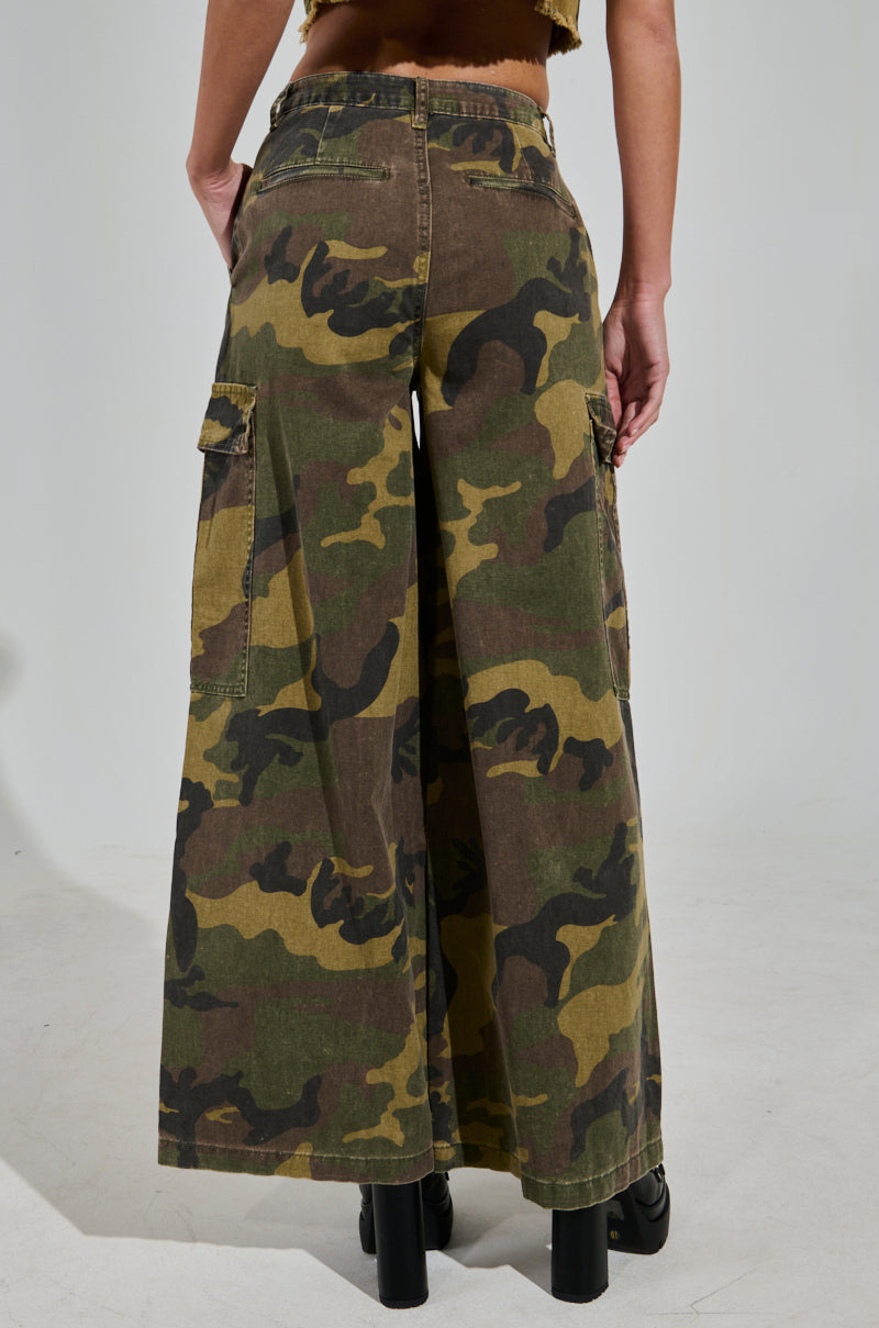 Someone is wearing the RIDE WITH ME CAMO WIDE LEG PANT, featuring high-rise and cargo-style pockets, paired with black platform shoes. The green and brown camouflage pattern stands out from the back view.