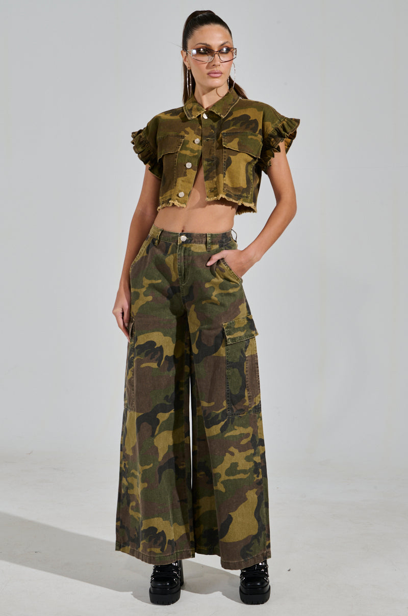 Dressed confidently in a cropped, frilled camo jacket and RIDE WITH ME CAMO WIDE LEG PANT featuring high-rise and cargo-style pockets, the individual sports sunglasses and black platform boots with one hand in their pocket against a plain backdrop.