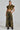 Dressed confidently in a cropped, frilled camo jacket and RIDE WITH ME CAMO WIDE LEG PANT featuring high-rise and cargo-style pockets, the individual sports sunglasses and black platform boots with one hand in their pocket against a plain backdrop.