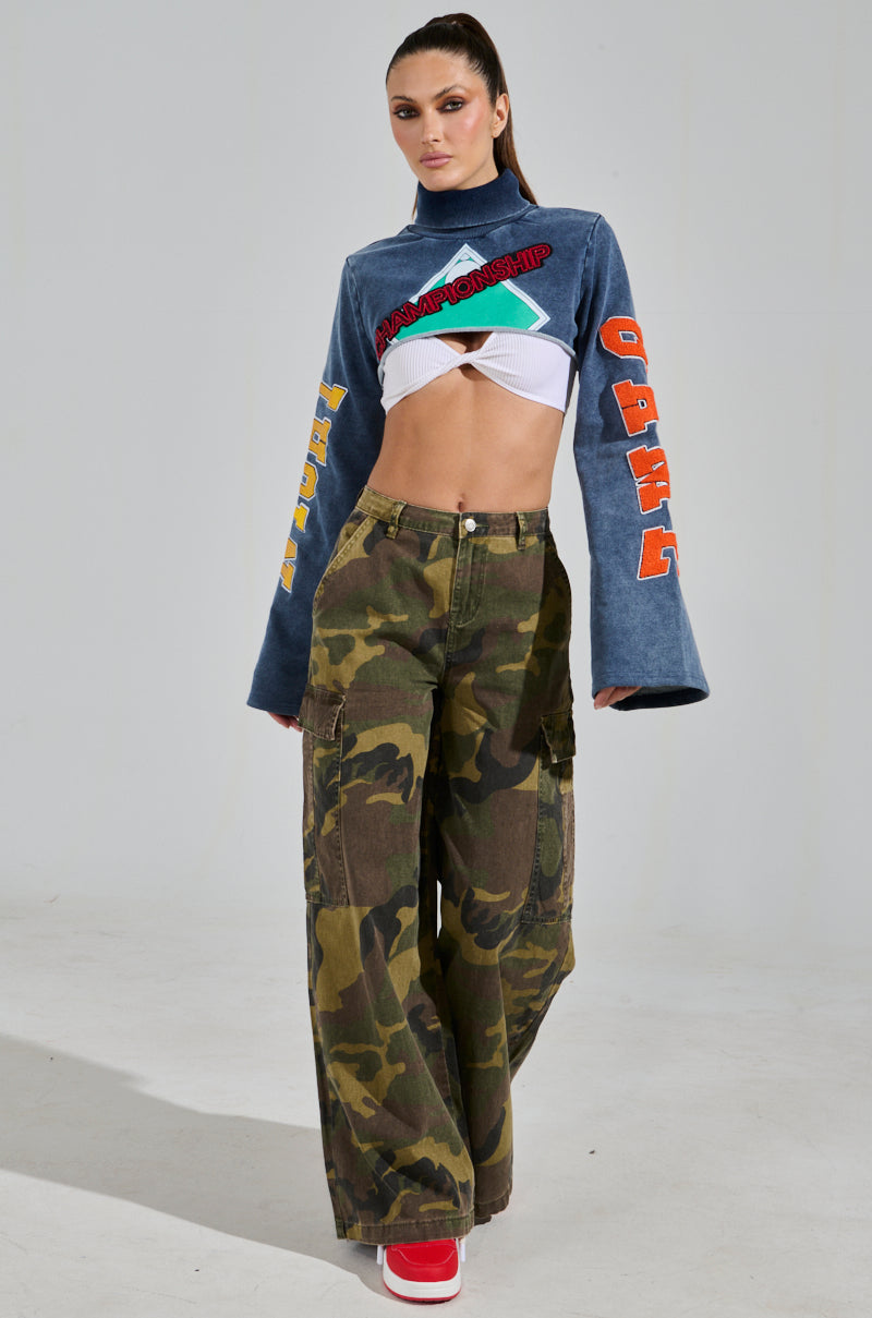 A person poses confidently against a light background, wearing a blue cropped top with colorful sleeve text and RIDE WITH ME CAMO WIDE LEG PANT featuring cargo pockets. They complete the outfit with bright red shoes and have a high ponytail.