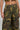The person models the RIDE WITH ME CAMO WIDE LEG PANT, highlighting its high-rise fit, camo pattern, wide-leg silhouette, cargo pockets, and button closure as they stand against a plain background with one hand on their hip.