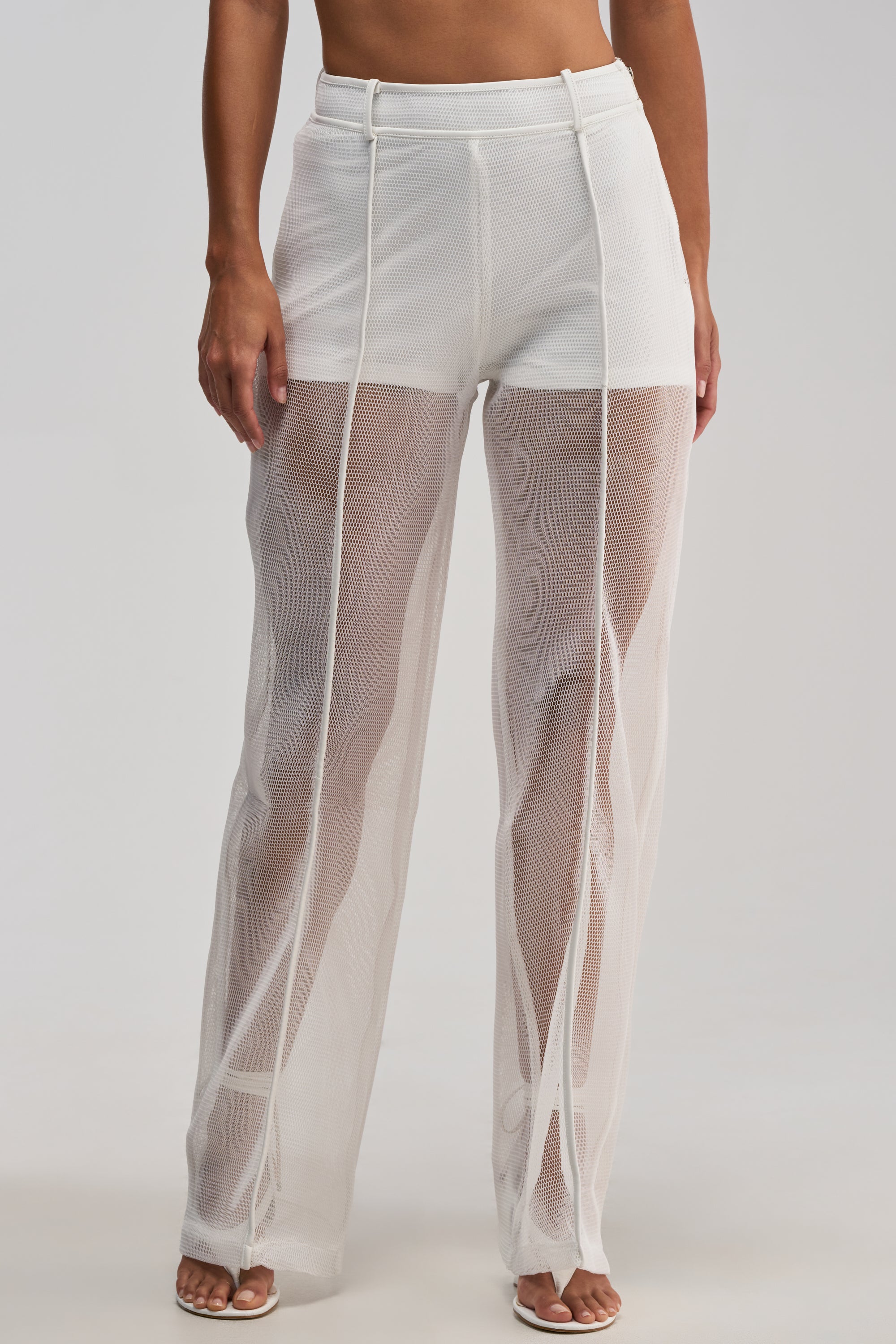 The model showcases the ONE TO WATCH MESH TROUSER IN WHITE—white sheer mesh wide-leg pants with drawstring detail—styled for a chic streetwear vibe and paired with white sandals against a gray background.