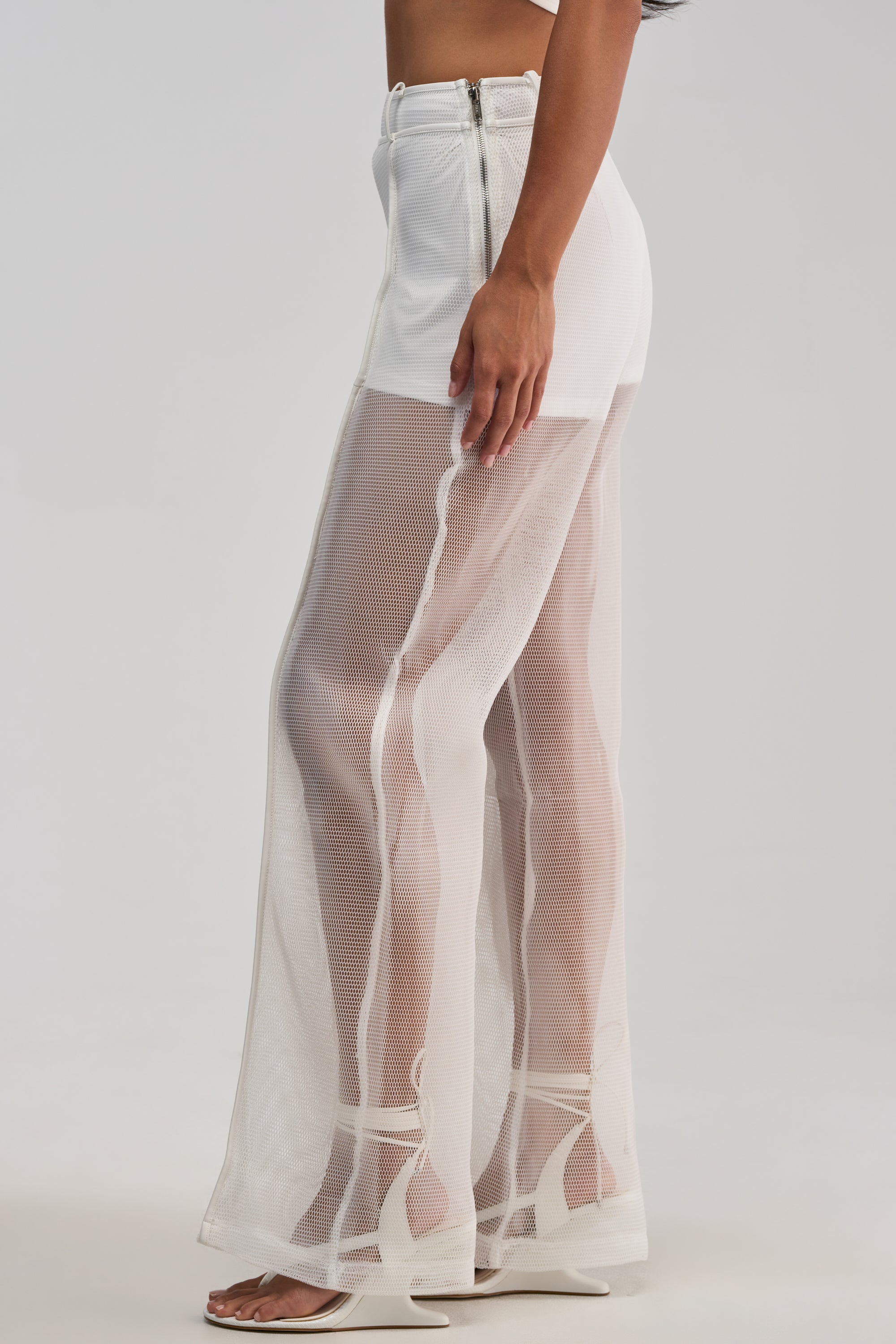 Styled for a modern summer look, she wears the ONE TO WATCH MESH TROUSER IN WHITE—high-waisted pants featuring sheer mesh fabric from mid-thigh down and a visible side zipper—paired with white sandals. Ideal for date nights or chic streetwear vibes, she stands sideways against a plain background.