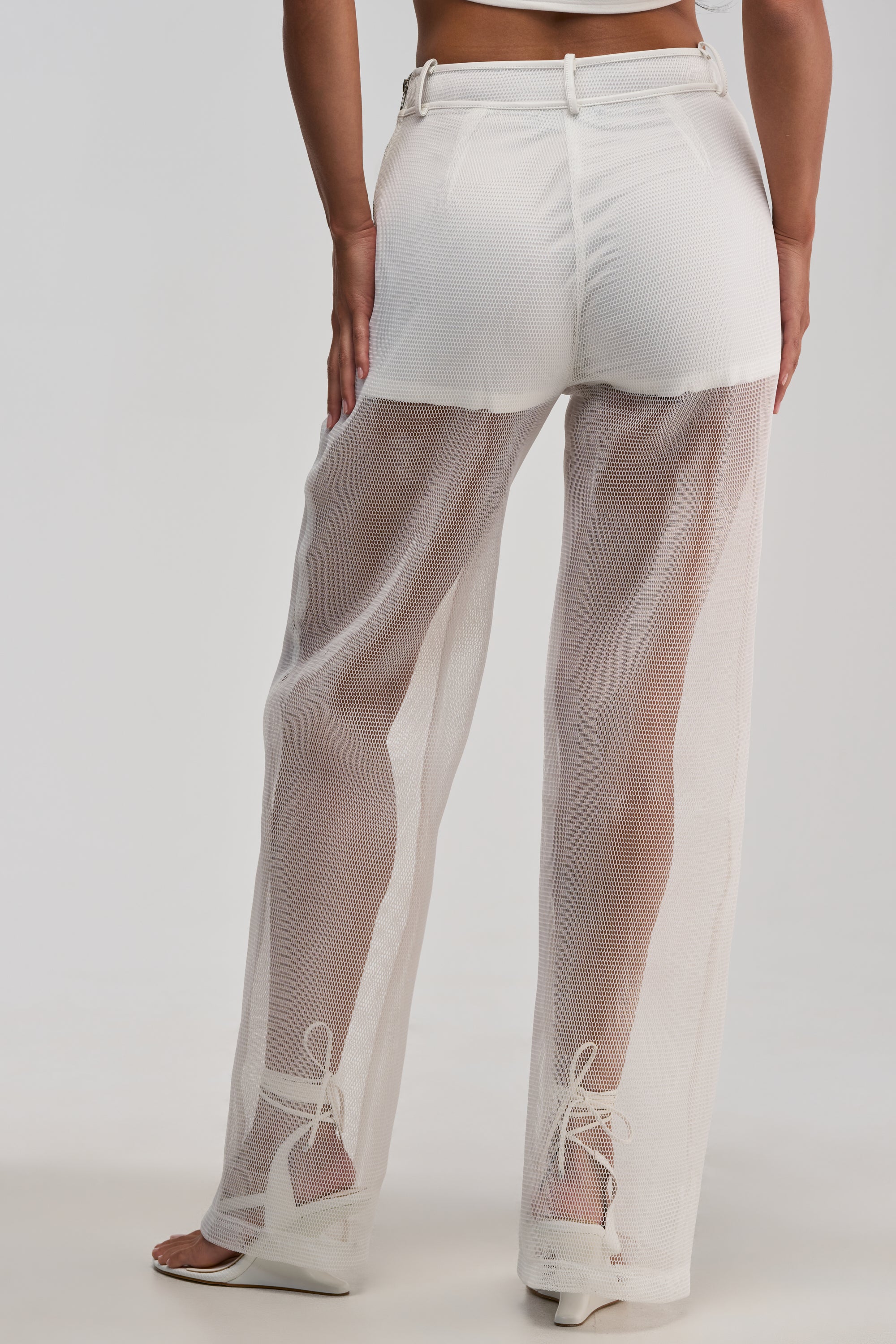 Model wearing the ONE TO WATCH MESH TROUSER IN WHITE, styled as high-waisted, sheer mesh pants layered over white underwear and paired with white sandal heels, shown from the back against a light background—a chic look ideal for streetwear styling or a summer date night.