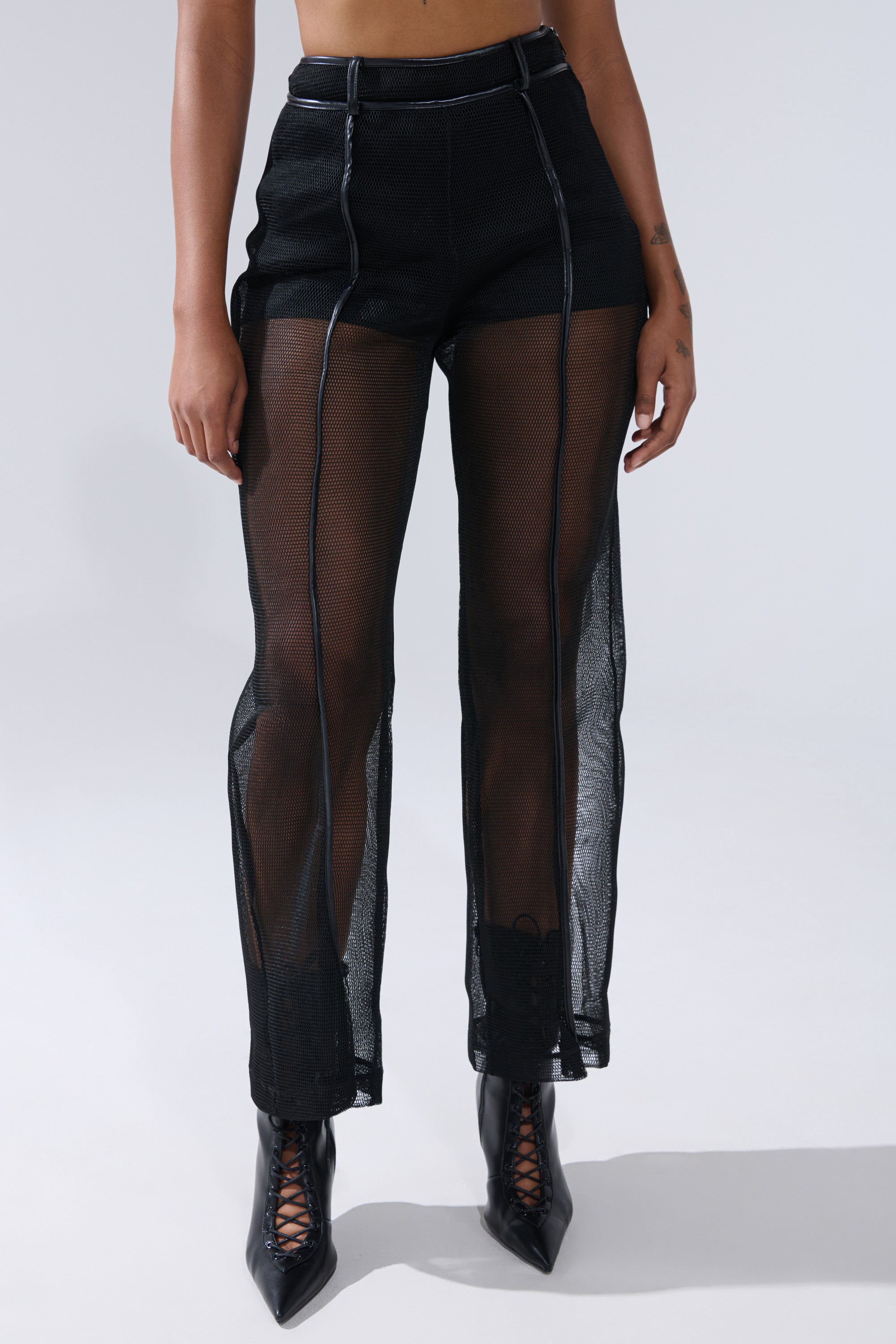 Wearing the ONE TO WATCH MESH TROUSER with high-rise fit, black mesh panels, and faux leather trim, paired with black lace-up ankle boots, a person stands against a plain light background. 0