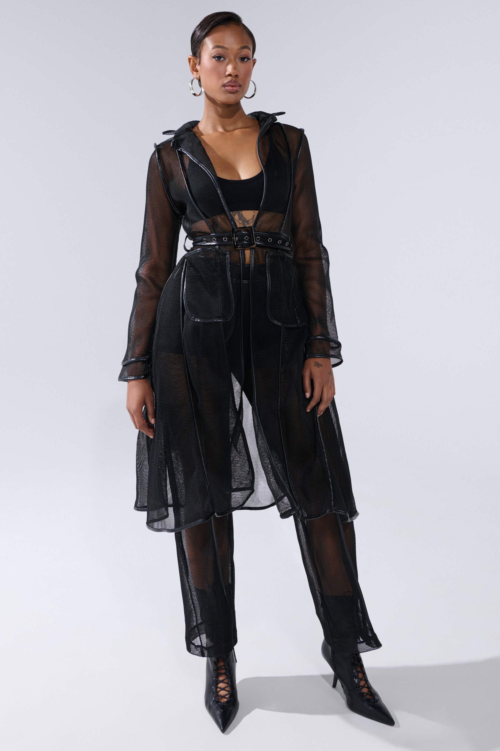 A woman poses confidently in the ONE TO WATCH MESH TROUSER, styled with a sheer black trench coat, matching crop top, and pointed lace-up heels. She wears hoop earrings and has her hair pulled back against a plain gray background. 5
