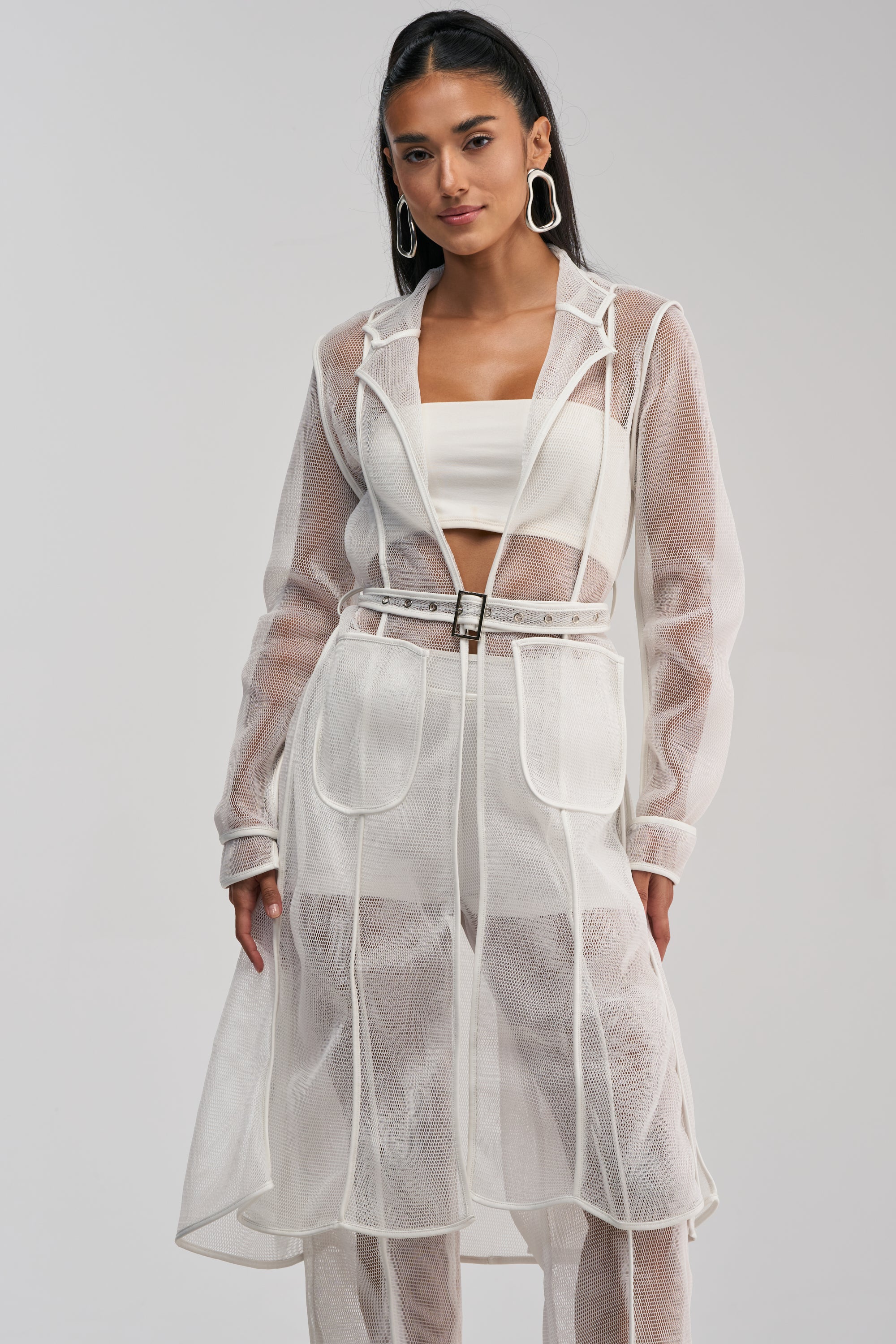 Wearing the ONE TO WATCH MESH TRENCH IN WHITE over a white crop top and pants, a woman with long dark hair and large geometric earrings perfectly embodies effortless summer streetwear against a plain light background.