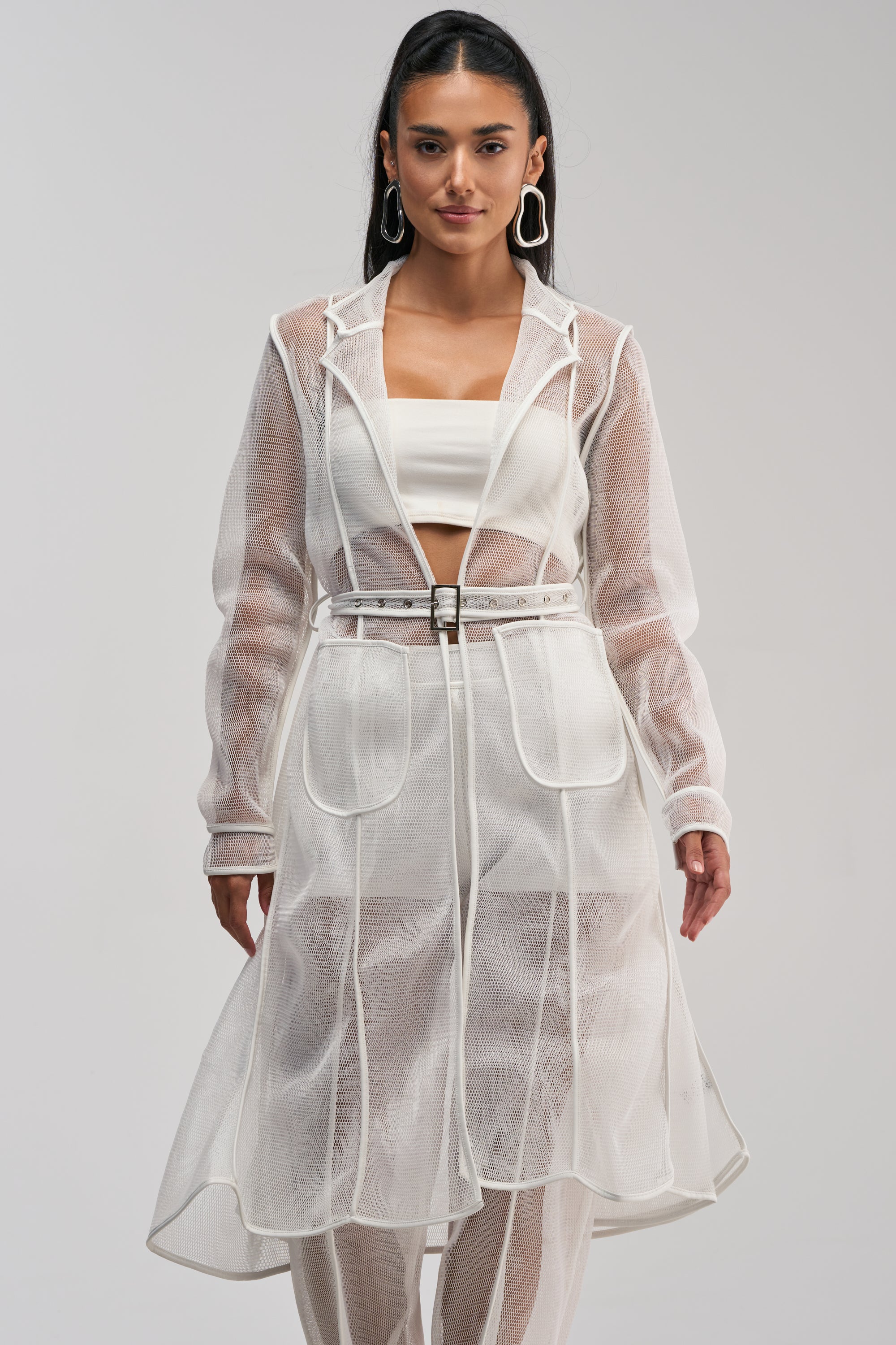 A woman wearing the ONE TO WATCH MESH TRENCH IN WHITE over a white bandeau top and matching pants, posing against a plain light background.
