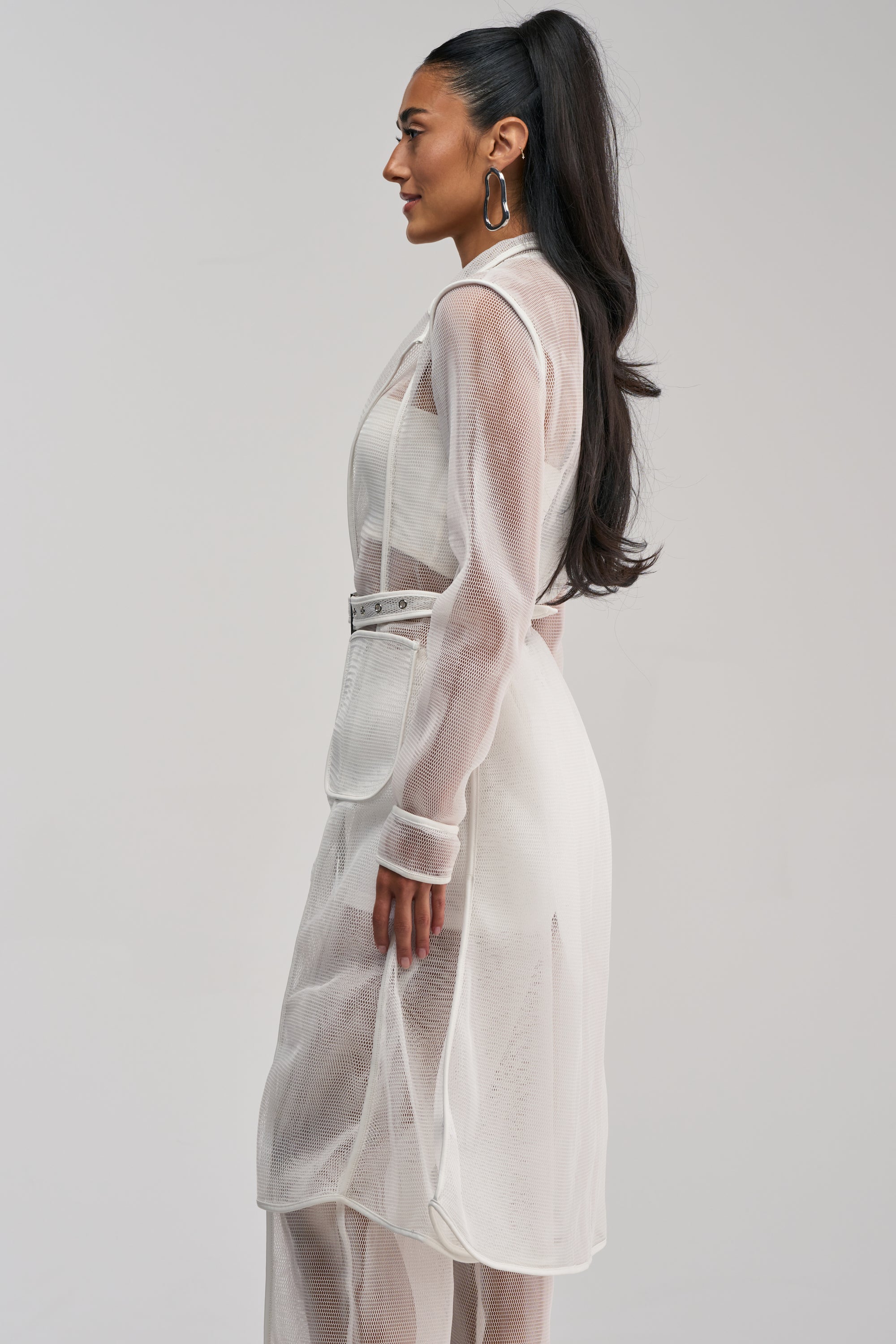 Wearing the ONE TO WATCH MESH TRENCH IN WHITE, this woman is styled in profile with long, dark, wavy hair pulled into a high ponytail and silver hoop earrings. She layers the sheer white long-sleeve trench, cinched at the waist with a belt, over white pants for an effortlessly chic summer streetwear look against a simple light background.