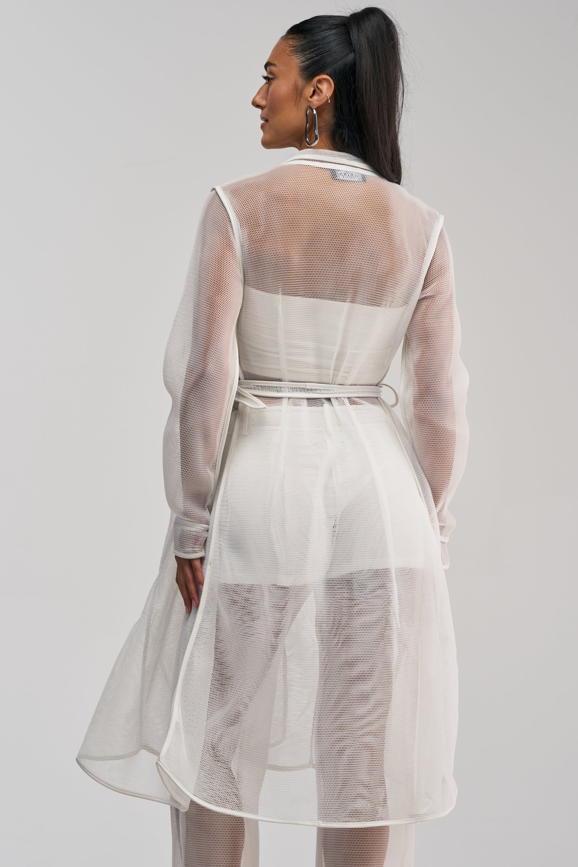 A woman with her dark hair styled in a high ponytail stands with her back to the camera, showcasing the ONE TO WATCH MESH TRENCH IN WHITE layered over a light outfit. The sheer, long-sleeved trench coat and minimalist light background perfectly evoke a modern summer streetwear aesthetic.