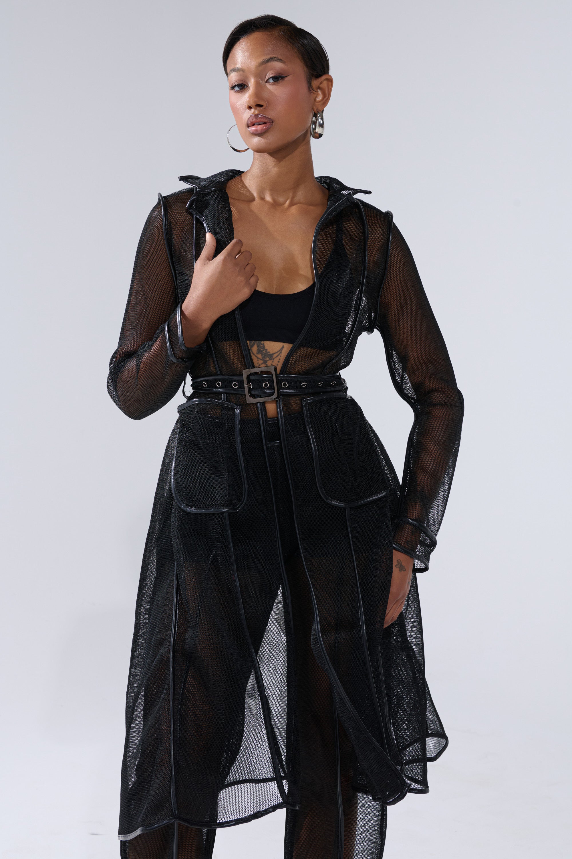 A woman poses confidently in the ONE TO WATCH MESH TRENCH IN BLACK with faux leather trim over a black outfit, hoop earrings, and short hair, standing against a plain light gray background. 0