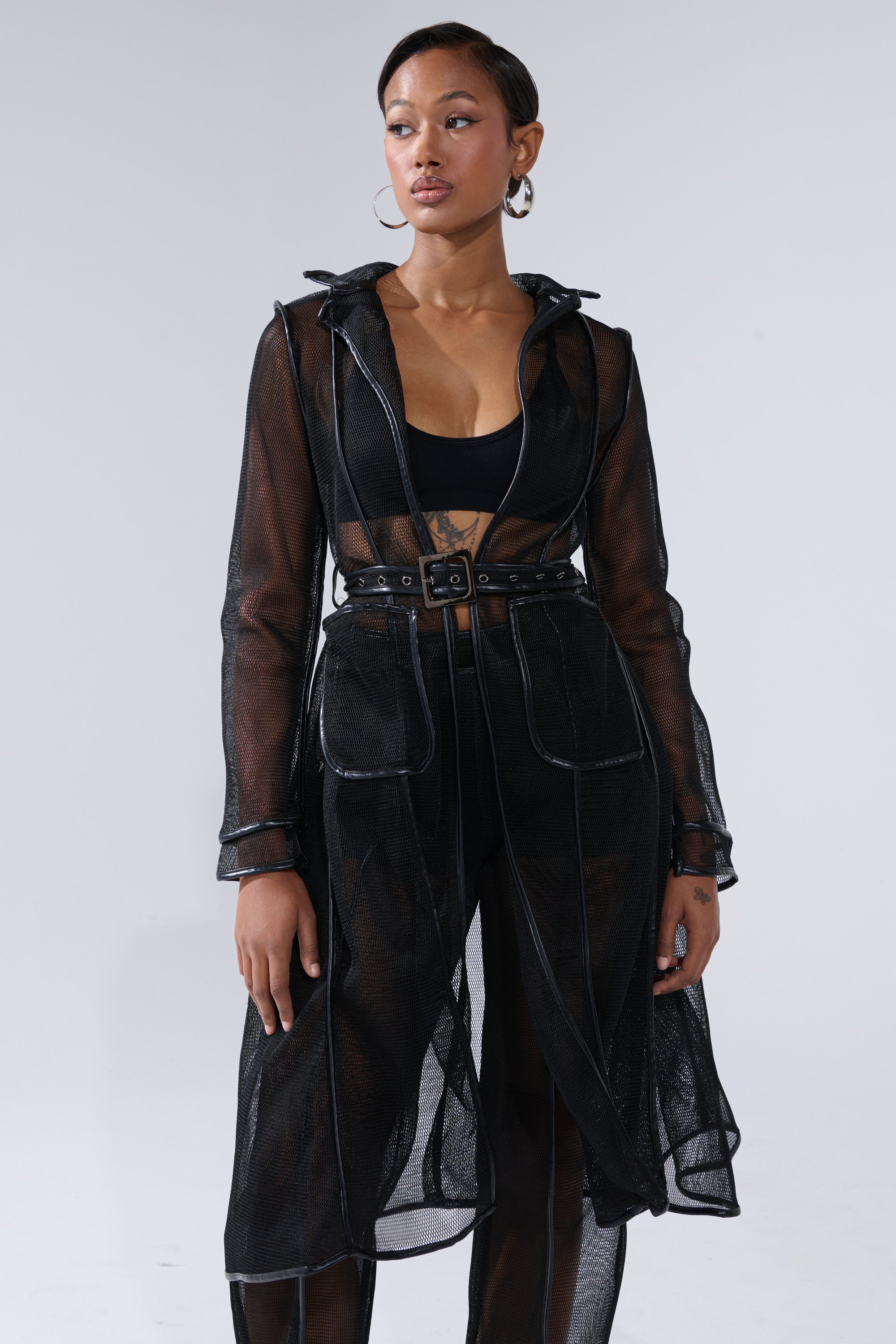 A woman wears the ONE TO WATCH MESH TRENCH IN BLACK over a black crop top and high-waisted pants, accessorized with hoop earrings, standing against a plain light gray background. 1