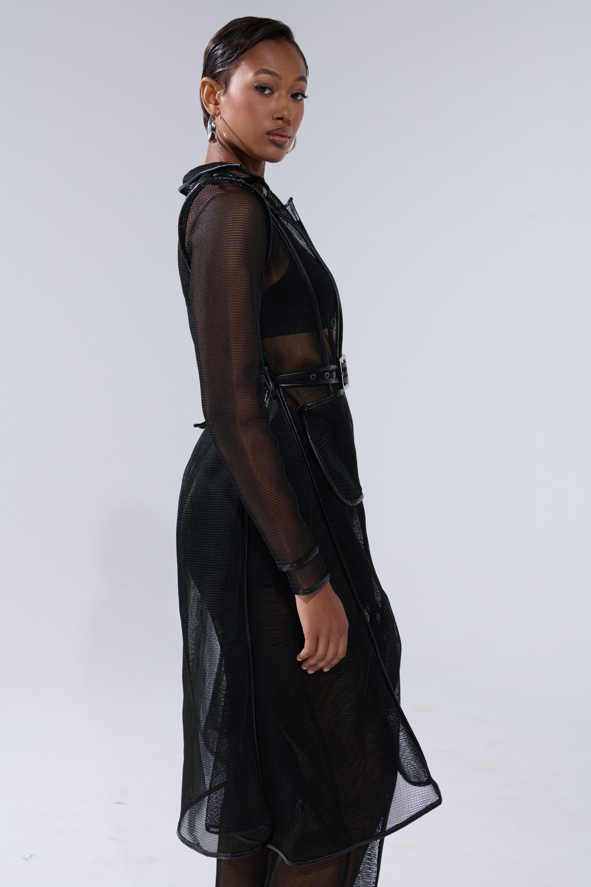 A woman stands in profile against a plain background, wearing the ONE TO WATCH MESH TRENCH IN BLACK, a sheer black long-sleeve trench with belted details and faux leather trim. She has short, slicked-back hair and a confident expression. 2