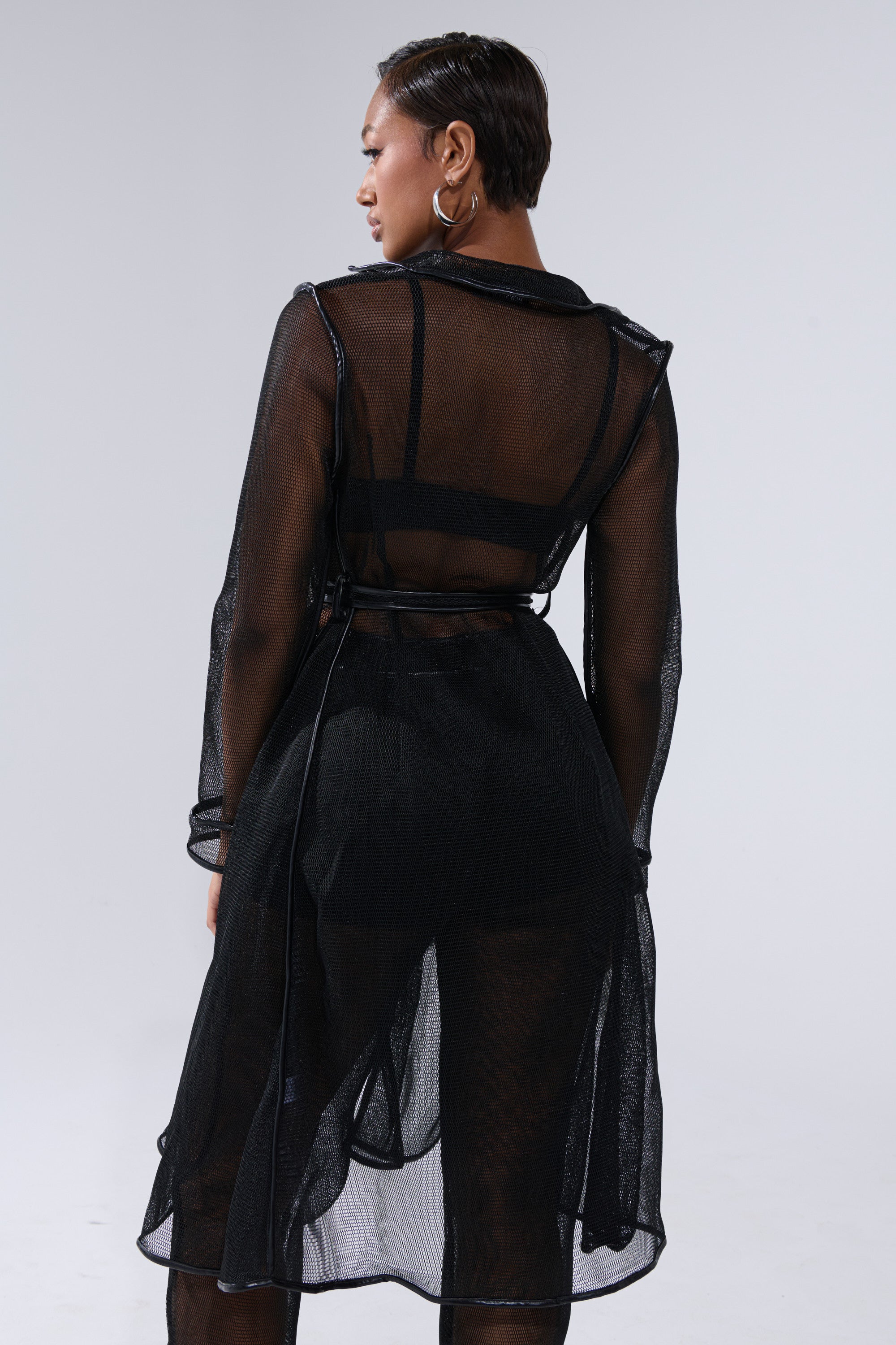 A person with short hair models the ONE TO WATCH MESH TRENCH IN BLACK over a black bra and shorts, standing with their back to the camera against a plain light gray background. 3