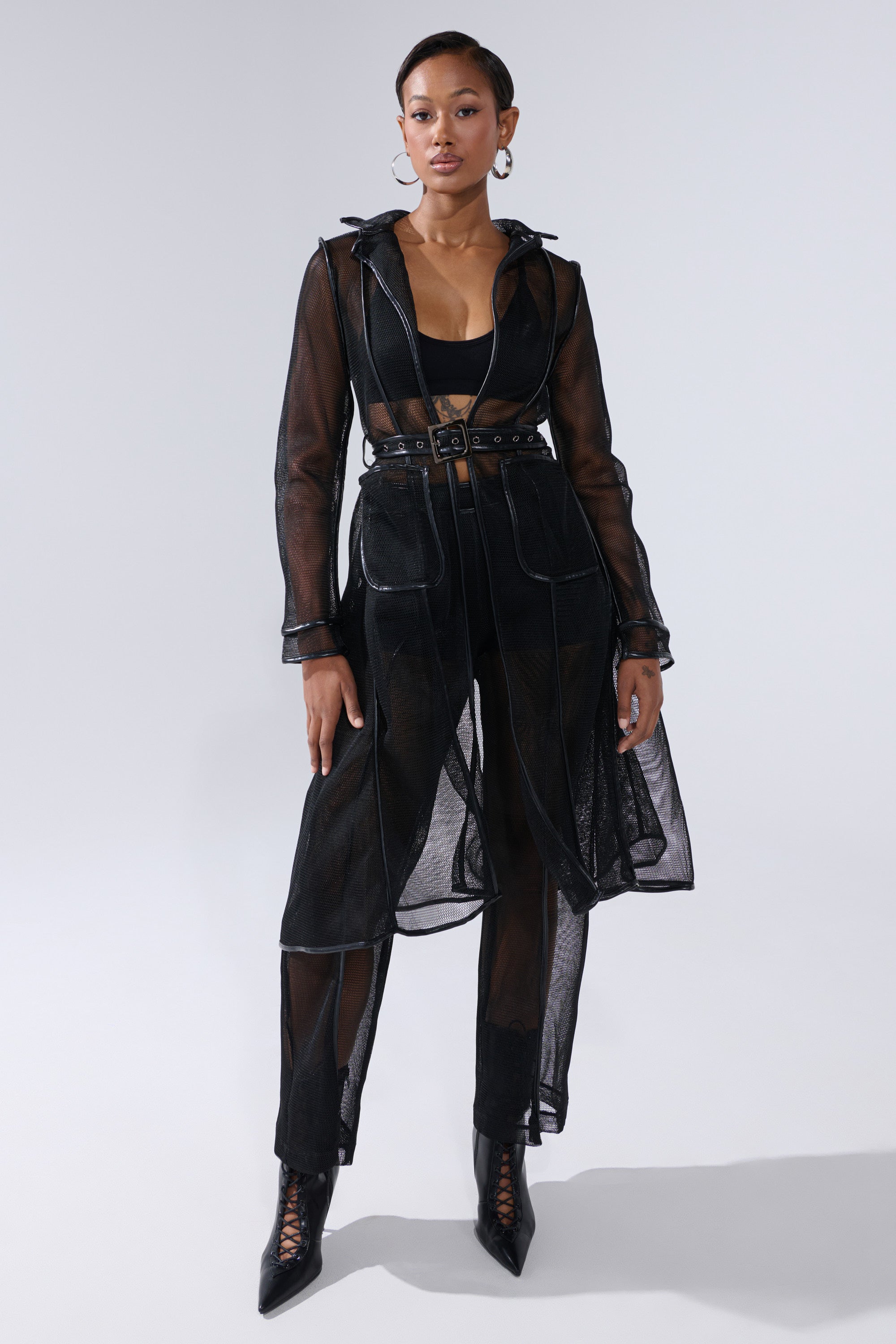 A woman exudes confidence in the ONE TO WATCH MESH TRENCH IN BLACK, layered over a black crop top and high-waisted pants with pointed lace-up boots. She accessorizes with hoop earrings and wears her hair pulled back. 4