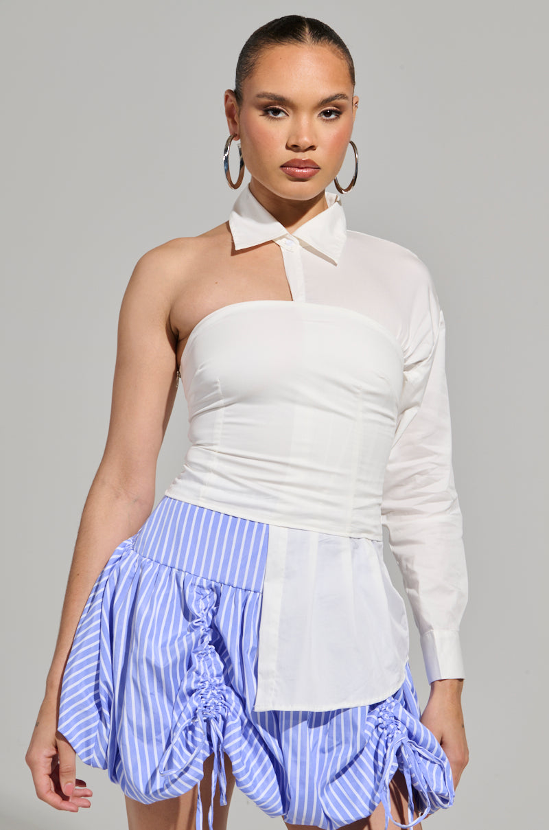 A person poses confidently against a gray background, showcasing a stylish ensemble that includes the "DONT LOOK AT ME ASYMMETRICAL POPLIN BLOUSE" with a one-sleeve design and a blue-striped skirt with ruching. Large hoop earrings complete the look, adding flair to their cotton blend top.