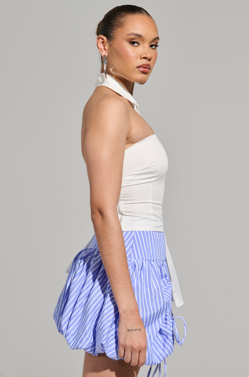 A person stands sideways, gazing forward in the DONT LOOK AT ME ASYMMETRICAL POPLIN BLOUSE layered over a blue and white striped skirt. Their hair is pulled back, and they sport hoop earrings. The neutral background enhances their chic ensemble.
