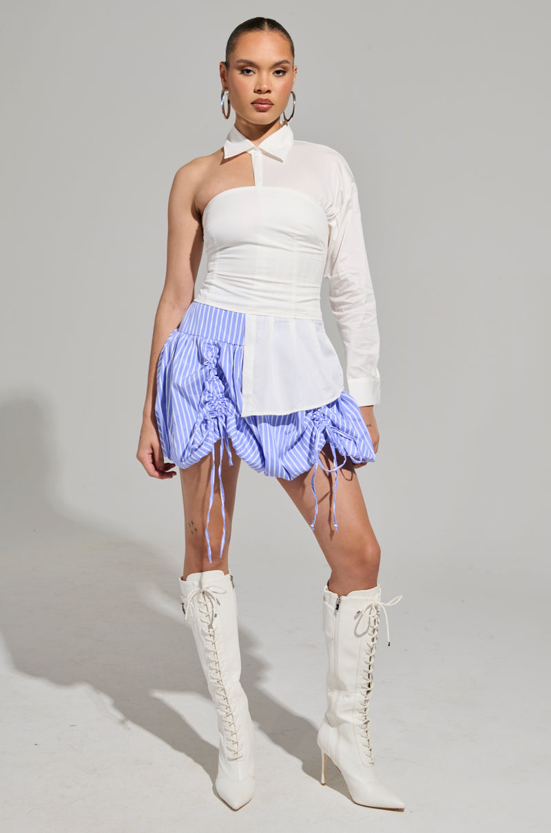 Wearing the DONT LOOK AT ME ASYMMETRICAL POPLIN BLOUSE with a chic blue-striped skirt featuring ruched detailing, the person confidently completes their look with knee-high white lace-up boots and large hoop earrings, set against a plain gray background.