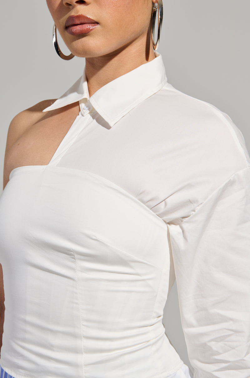 A person dressed in the DONT LOOK AT ME ASYMMETRICAL POPLIN BLOUSE, showcasing its distinct one-sleeve and cut-out design, complements their look with large hoop earrings. The plain, light-colored background accentuates the fashion-forward appeal of this trendy cotton blend blouse.