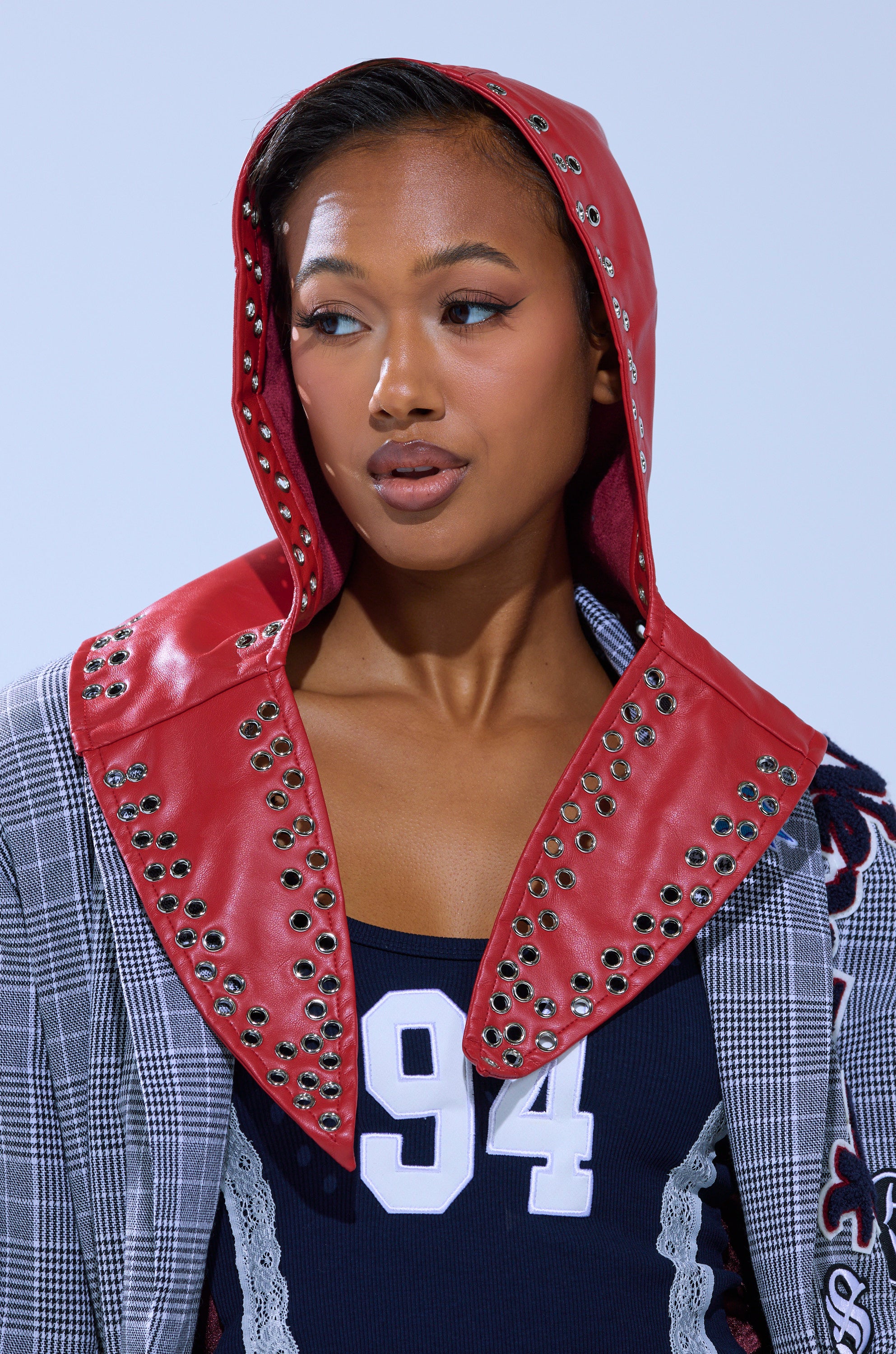 A woman wears the BUCKLE UP PU HEADSCARF—an oversized red studded faux leather hood—styled with a plaid jacket and a navy 94 top, looking to the side against a light blue background for a bold, standout look.