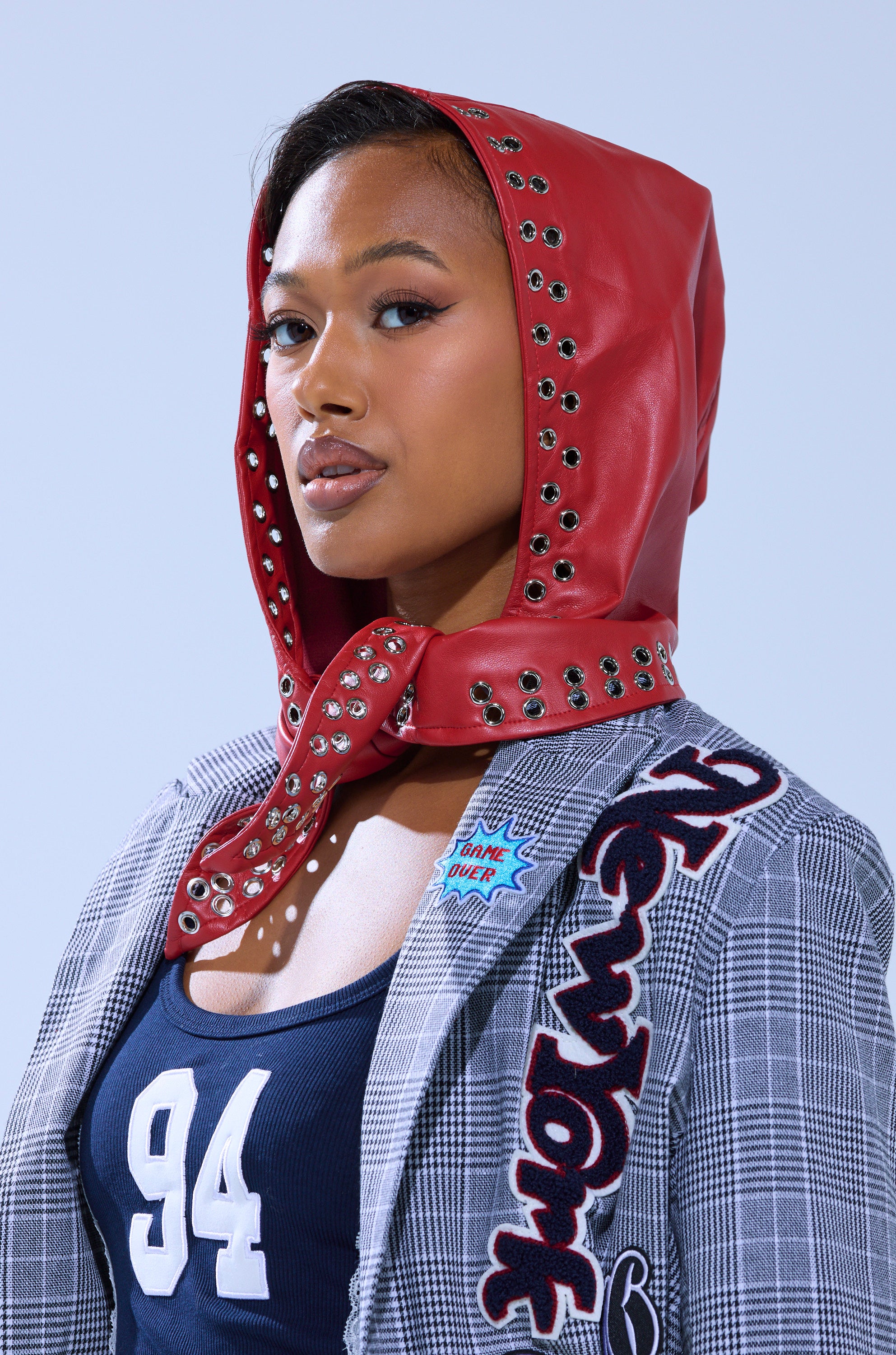 A person wearing the BUCKLE UP PU HEADSCARF—a bold red faux leather hood with metal eyelets—paired with a gray plaid blazer featuring "New York" lettering, patches, and a navy blue shirt with the number 94.