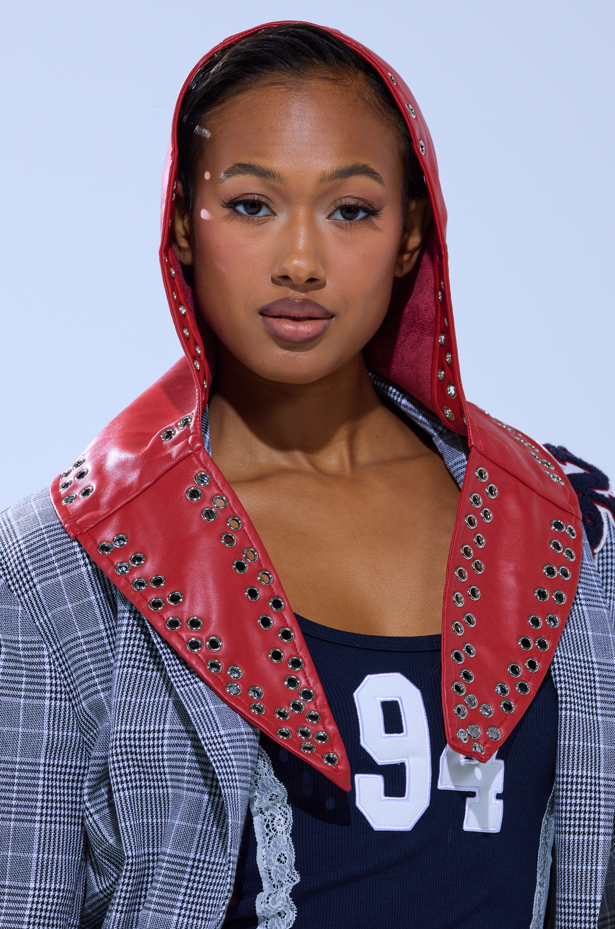 A woman wears the BUCKLE UP PU HEADSCARF—a red studded faux leather accessory—over a plaid jacket and navy blue top with the number 94, sporting white decorative face makeup near her eyes and looking confidently at the camera.