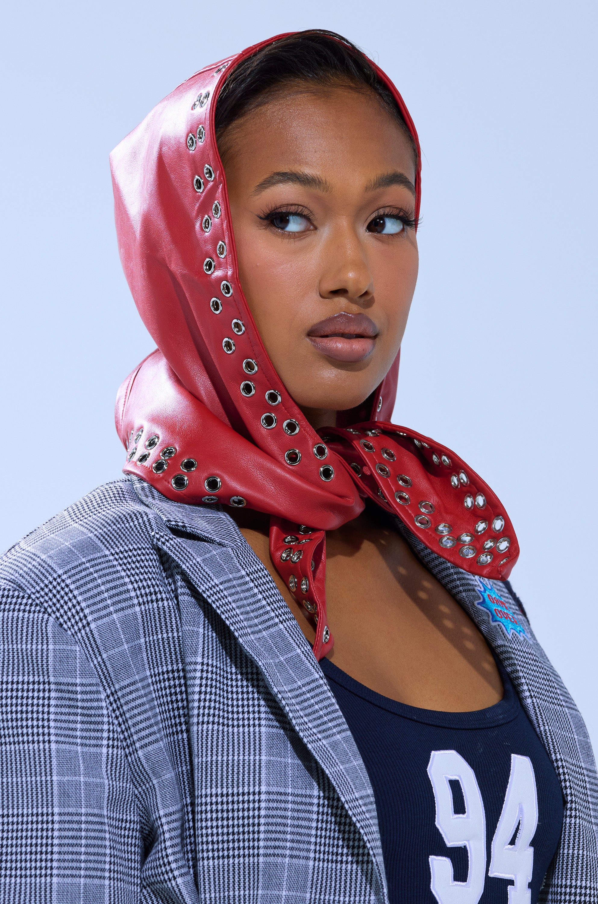 A woman wears the BUCKLE UP PU HEADSCARF in red with silver eyelets, styled with a black and white plaid blazer and navy blue top (No. 94), as she gazes to the side against a light blue backdrop.