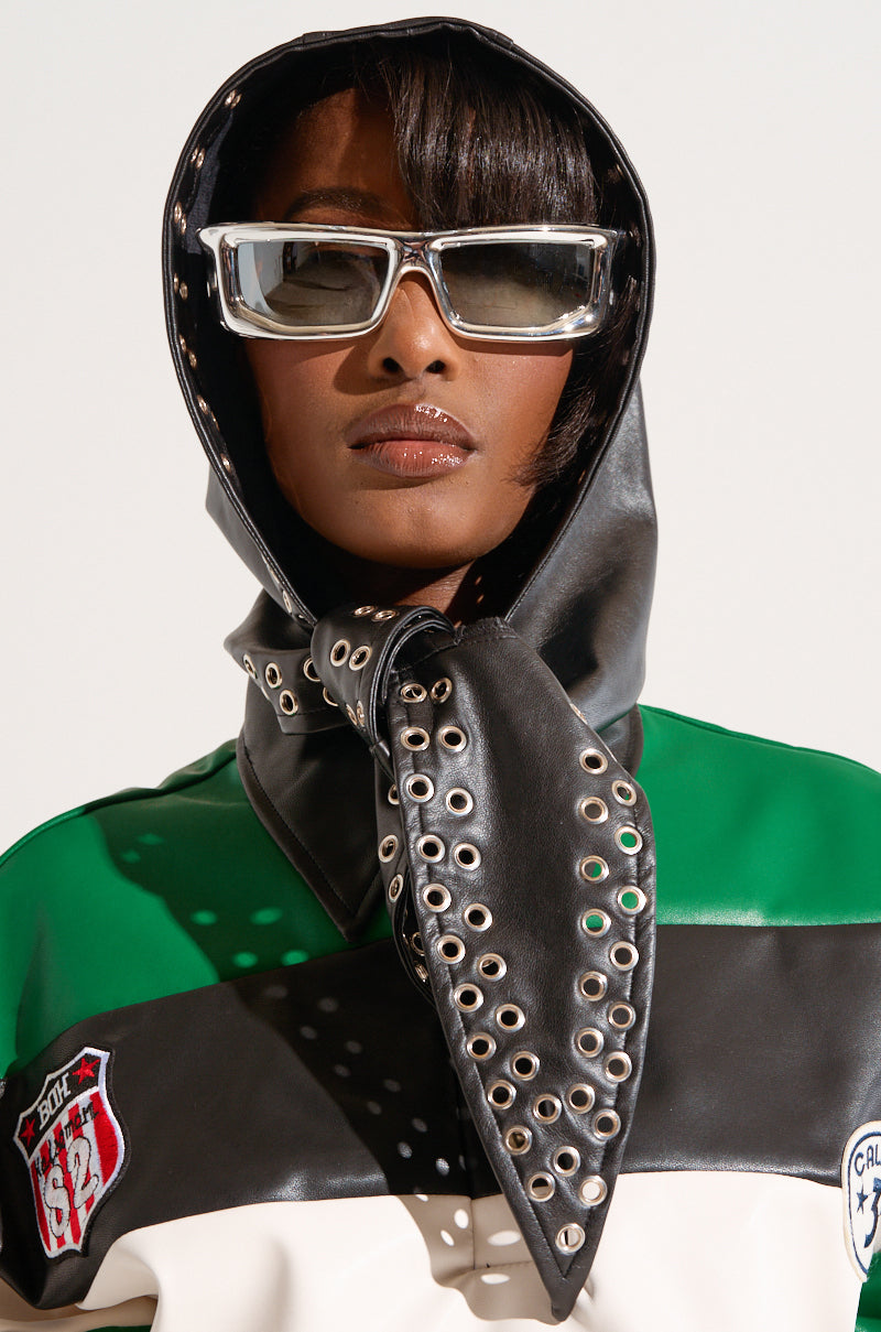 A person wearing the OH SO CHIC PU Head Scarf with metal grommets, white glasses, and a green, black, and white jacket adorned with patches. The background is minimal and light-colored, emphasizing the fashion-forward and edgy outfit.