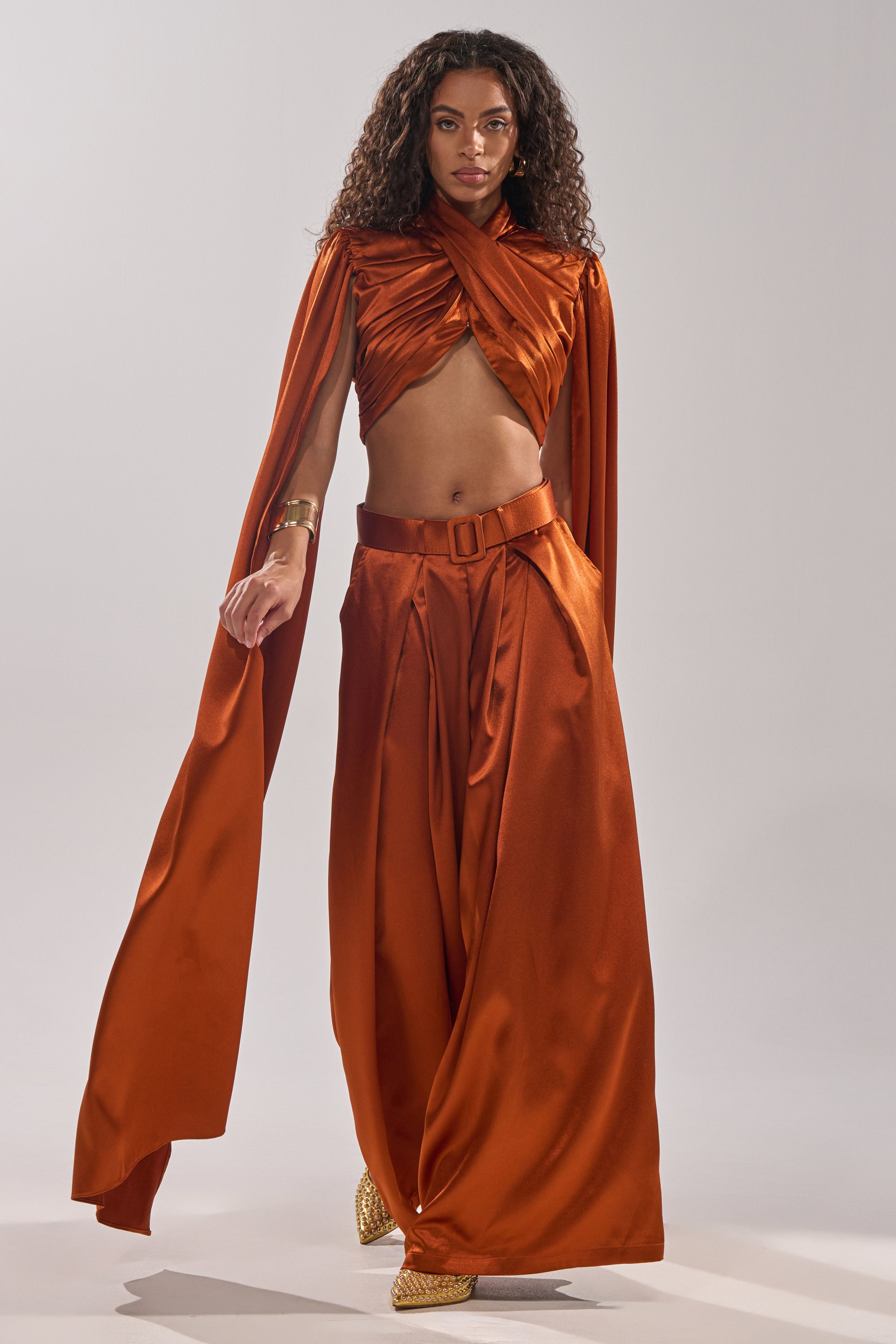 A woman wears the ALL WE EVER KNEW TROUSER, wide-leg satin pants in burnt orange with a high-rise fit, paired with a matching draped crop top and cape. She accessorizes with gold shoes and jewelry, her curly hair styled naturally against a light background.