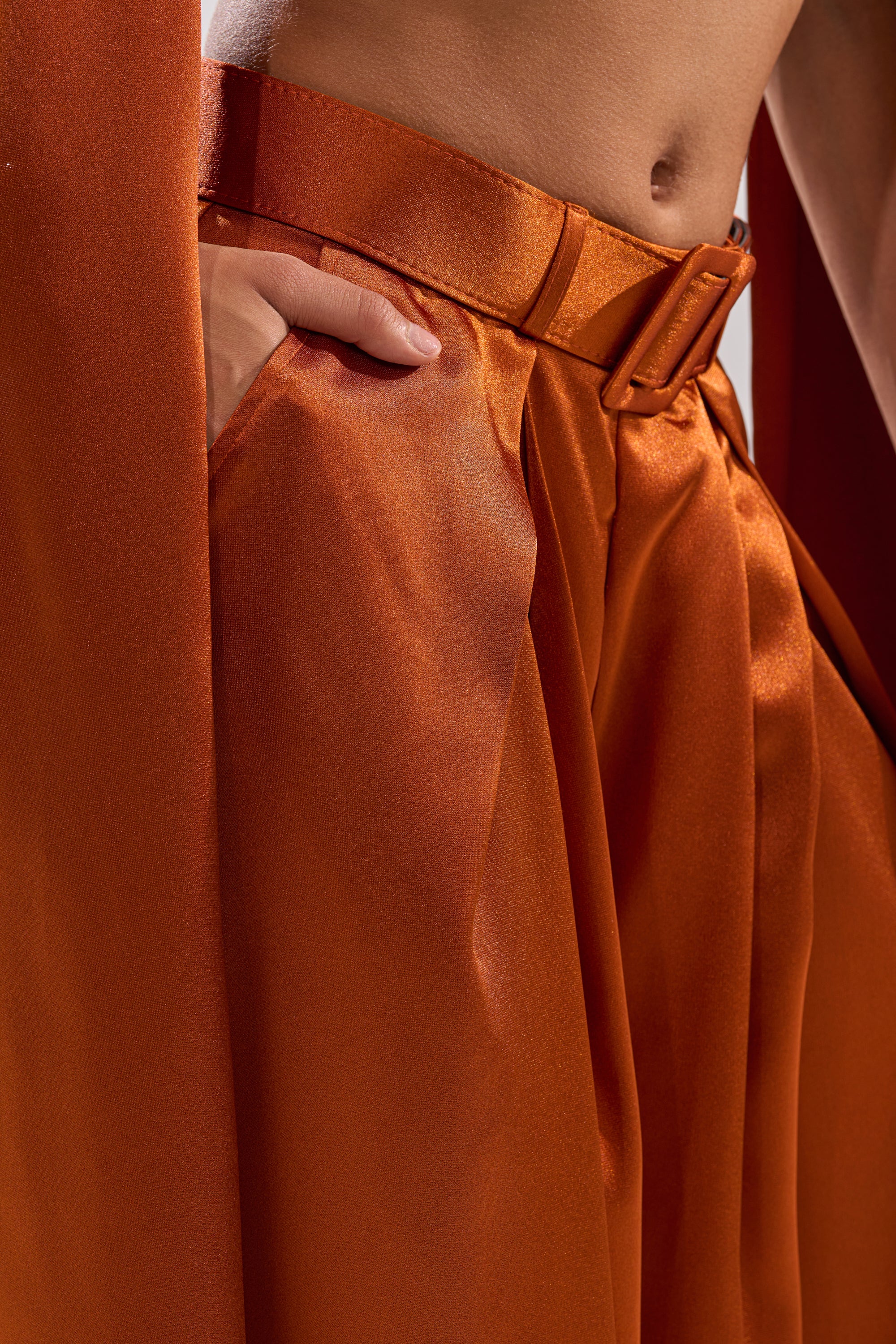 A close-up of someone wearing the ALL WE EVER KNEW TROUSER—shiny, rust-orange satin pants with a high-rise fit, pleats, matching belt, and one hand in the pocket. The smooth fabric has a silky appearance.