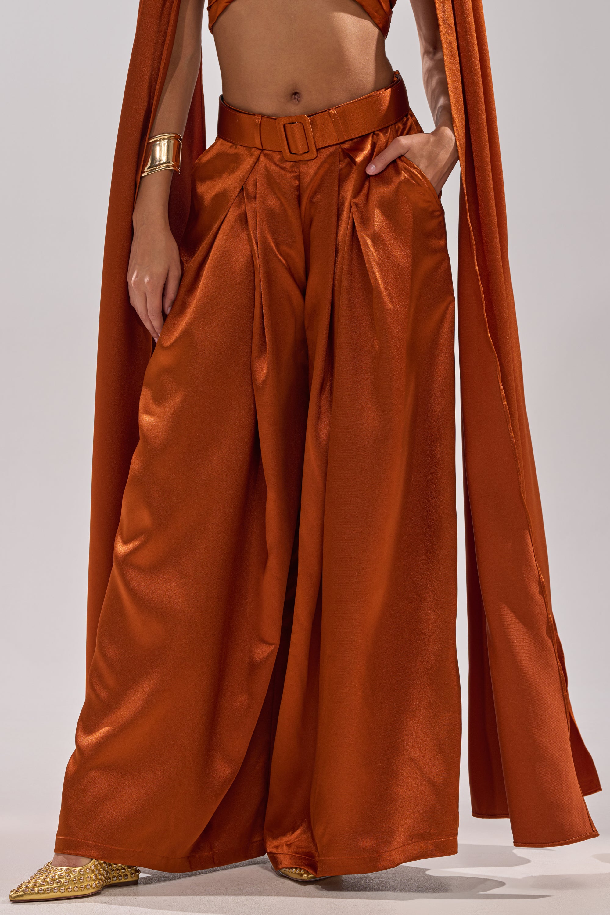 Wearing the ALL WE EVER KNEW TROUSER in burnt orange satin with a high-rise fit, wide-leg silhouette, matching belt and cape, this person accessorizes with a gold bracelet and pointed yellow shoes, hand casually in their pocket.