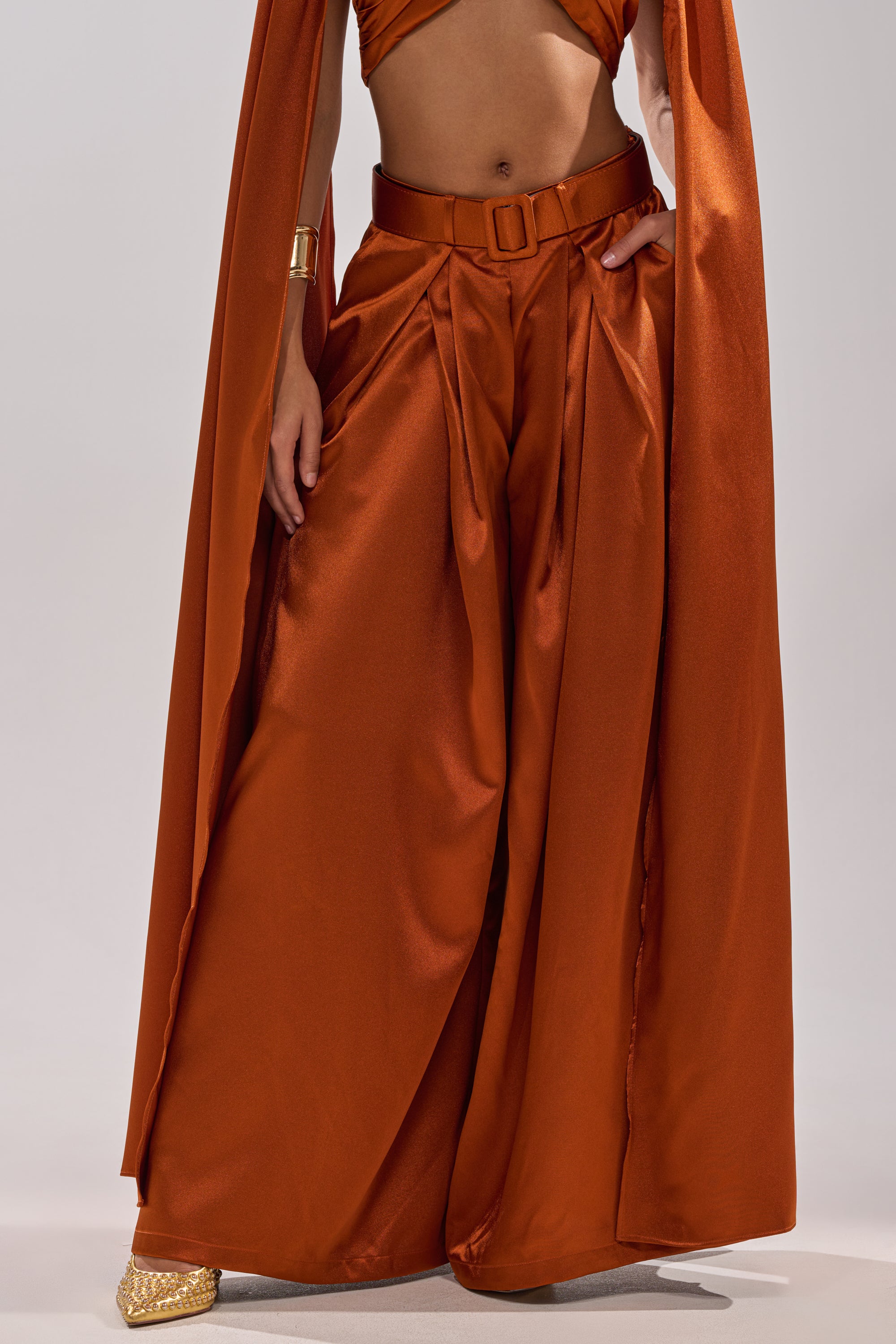 A person wears the ALL WE EVER KNEW TROUSER in rust-orange satin with a wide-leg, high-rise fit, styled with a coordinating top, cape, and gold pointed-toe shoes. Only the lower half of their body is visible.