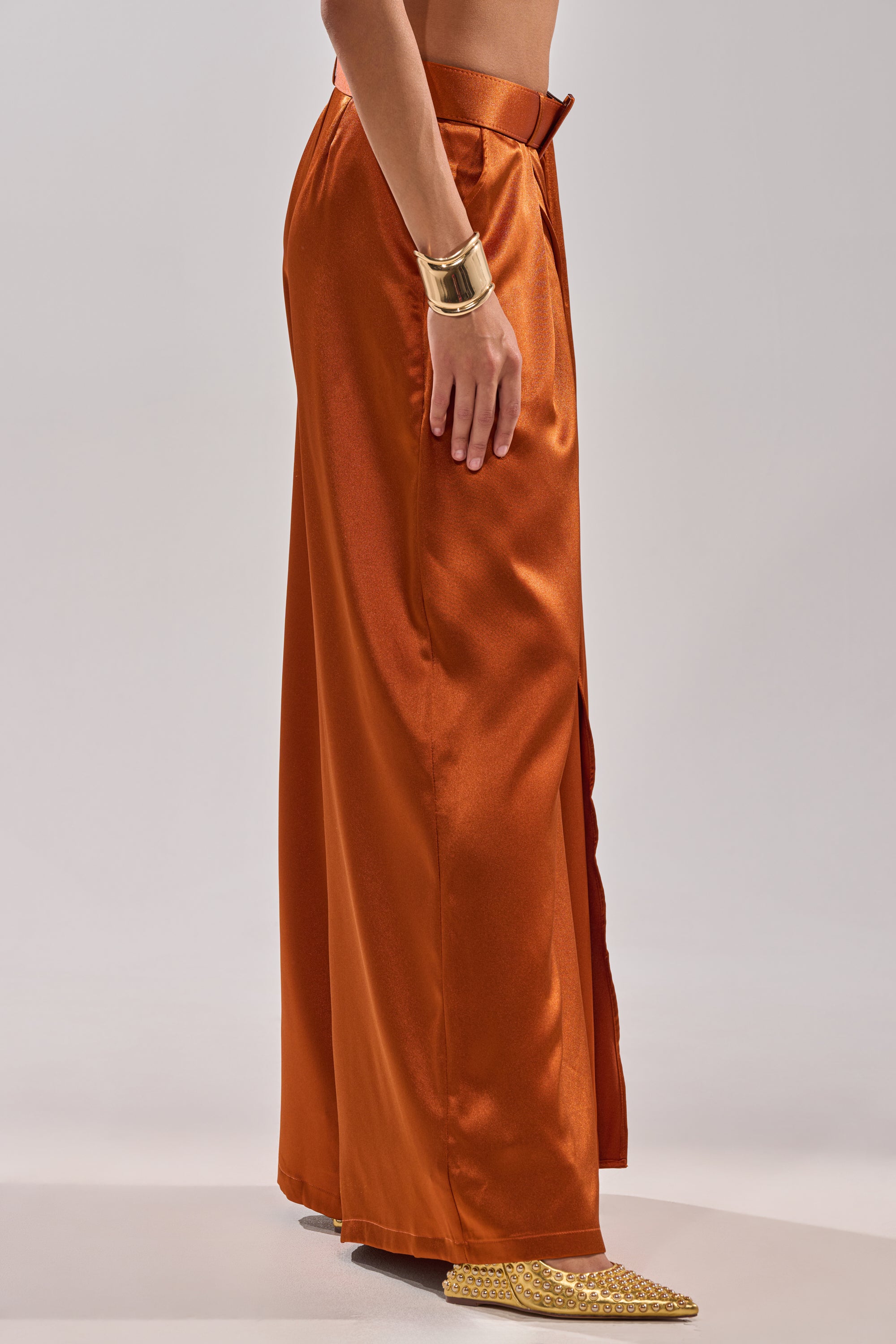 A model wears the ALL WE EVER KNEW TROUSER—shiny rust-orange satin, wide-leg with side slit—styled with a gold cuff bracelet and pointed gold shoes, standing against a plain light gray background.