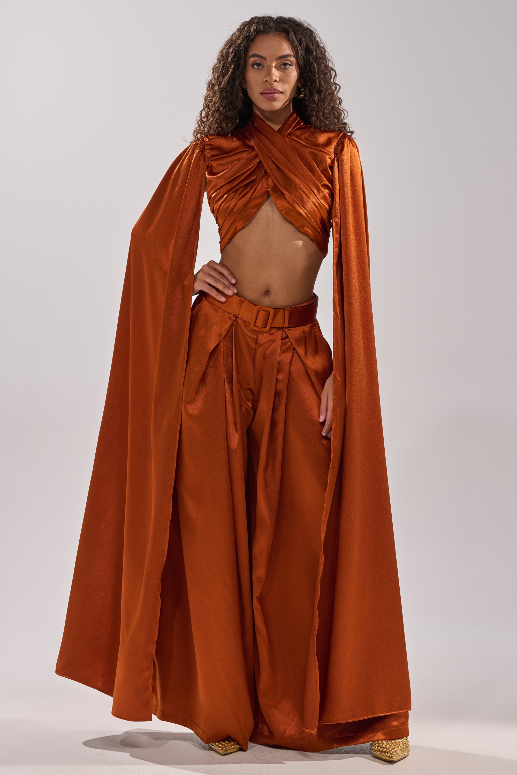 A woman with curly hair stands confidently against a plain background in the ALL WE EVER KNEW TROUSER—burnt orange, satin, high-rise wide-leg pants—styled with a draped crop top and matching floor-length cape, paired with gold shoes.