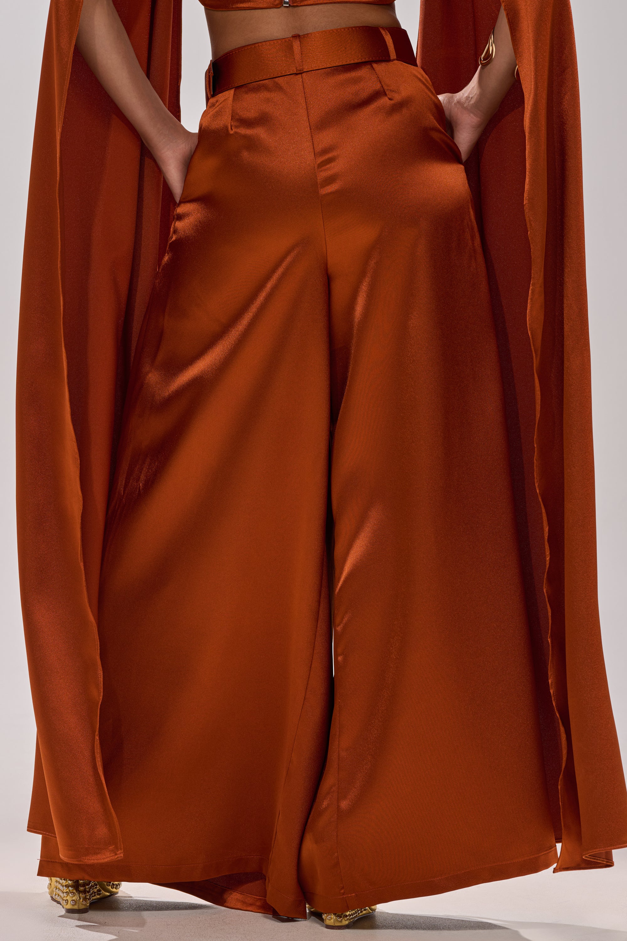 A person wears the ALL WE EVER KNEW TROUSER: shiny rust satin, wide-leg silhouette, flowing panels draped from a high-rise waist, standing with hands in pockets.