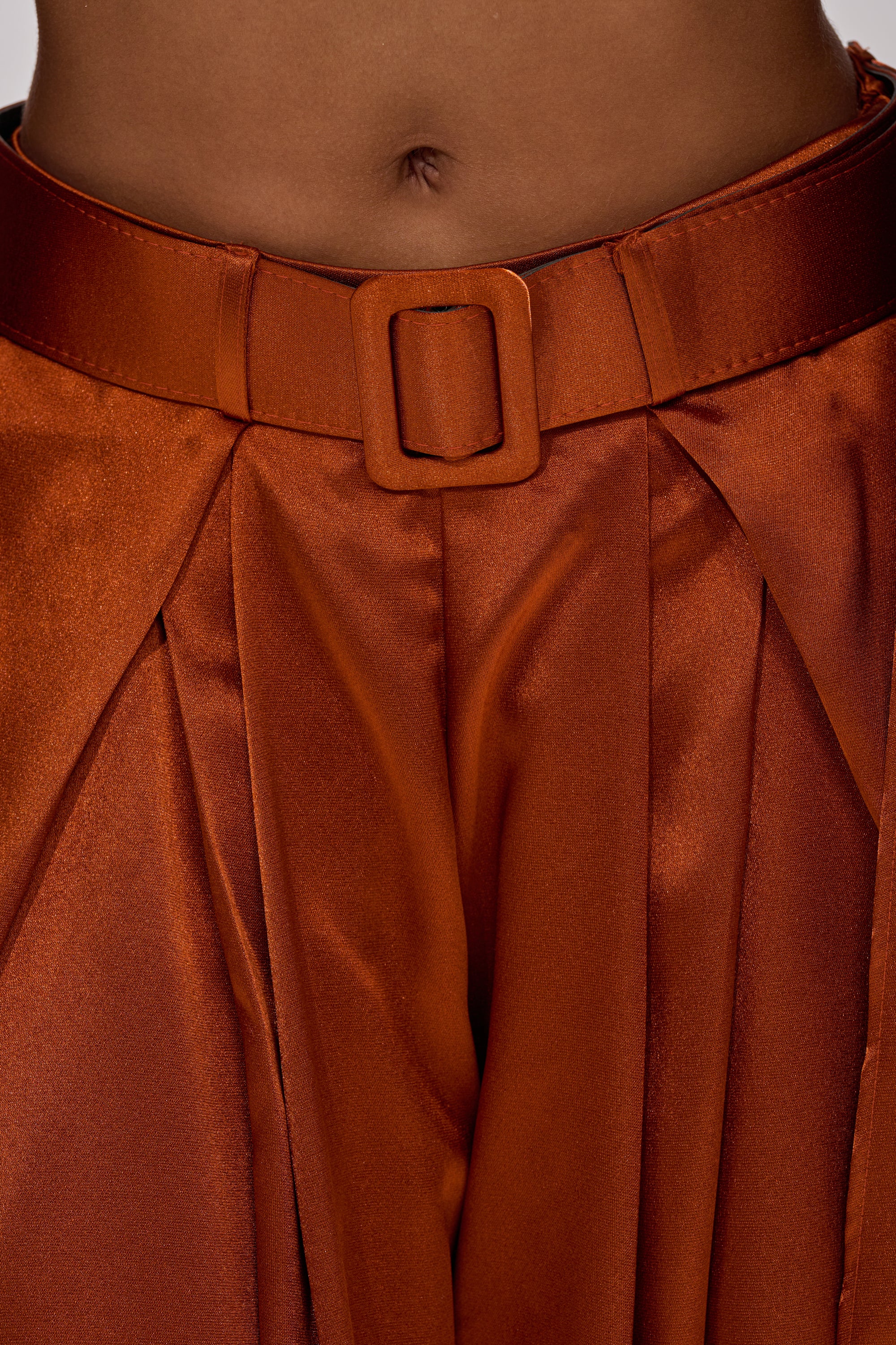 Close-up of a person wearing the ALL WE EVER KNEW TROUSER—shiny rust satin, high-rise fit with pleats, a wide-leg silhouette, and a matching belt with square buckle for added sophistication.