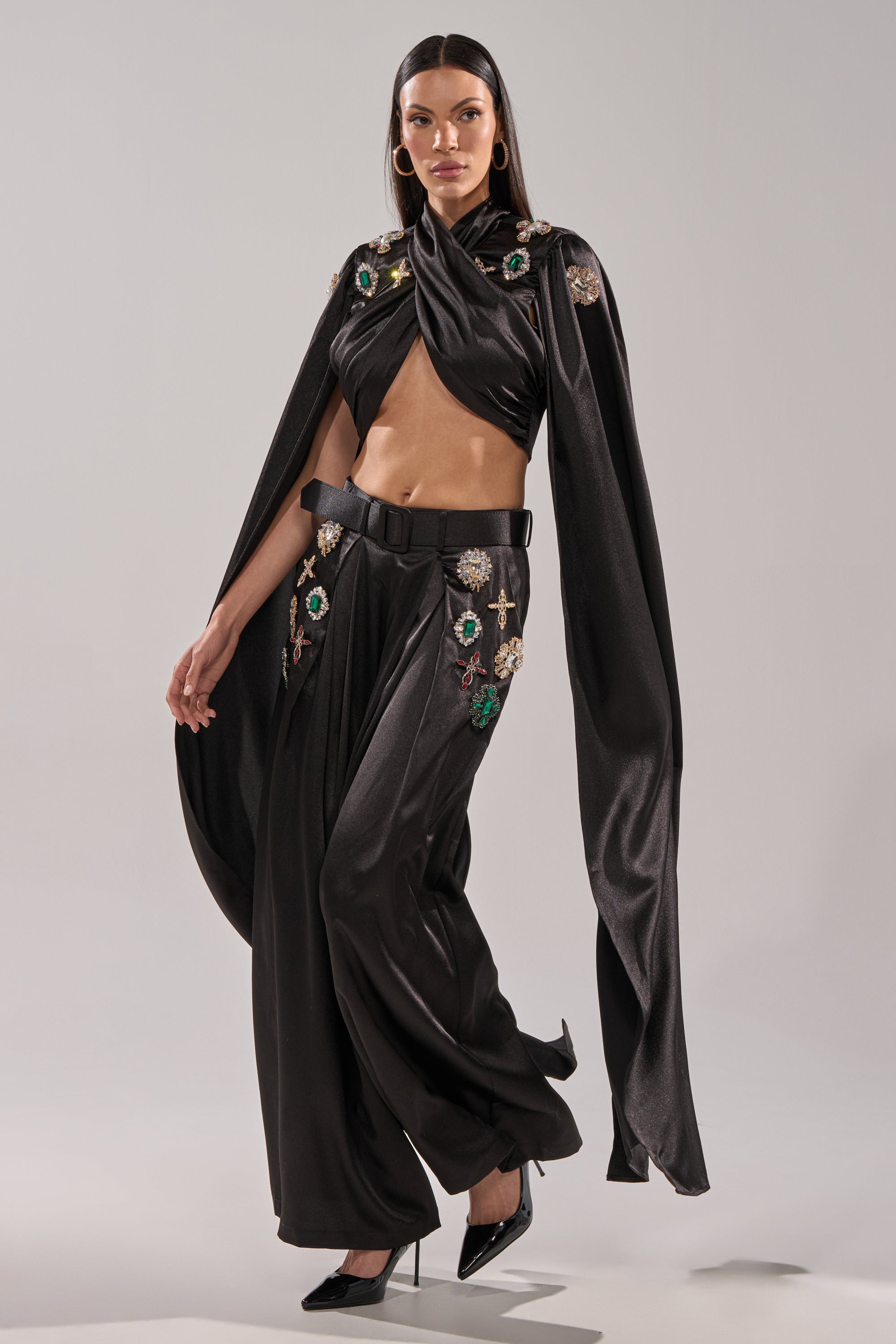 A woman stands against a plain light background in a black, shiny draped cape top and the AFTER DARK TROUSER—satin wide-leg pants featuring rhinestone brooch details—styled with a black belt and pointed heels.