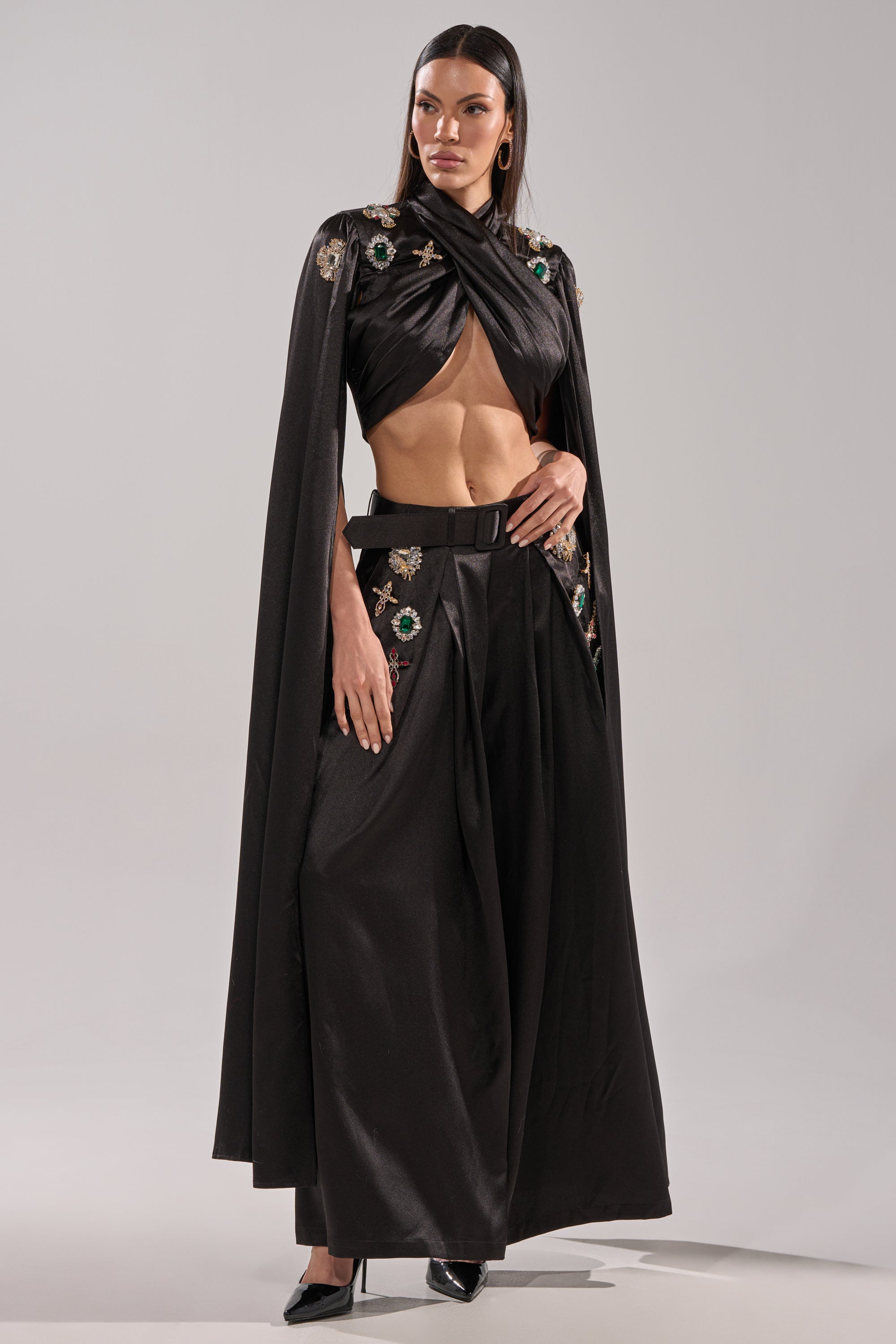 A woman stands confidently in the AFTER DARK TROUSER, paired with a fitted crop top and draped cape featuring floral embroidery and rhinestones. She has straight dark hair and finishes the look with black heels.