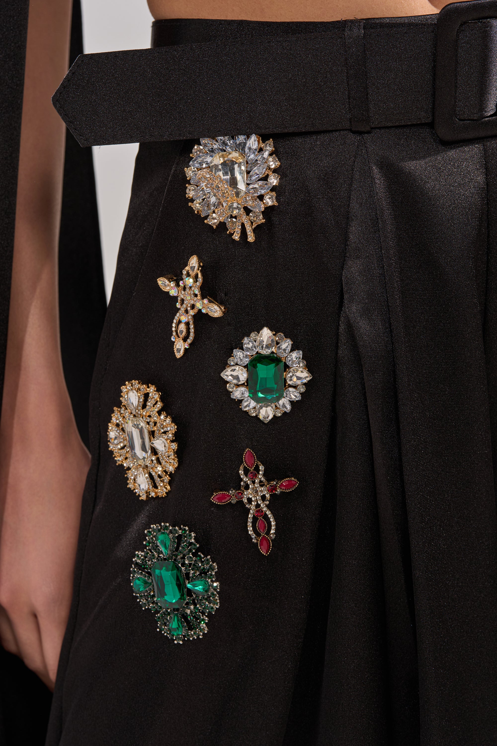 A close-up of a person wearing the AFTER DARK TROUSER: black pleated pants with a belt, embellished with green, red, and clear rhinestone brooches for bold sparkle on the wide-leg silhouette.