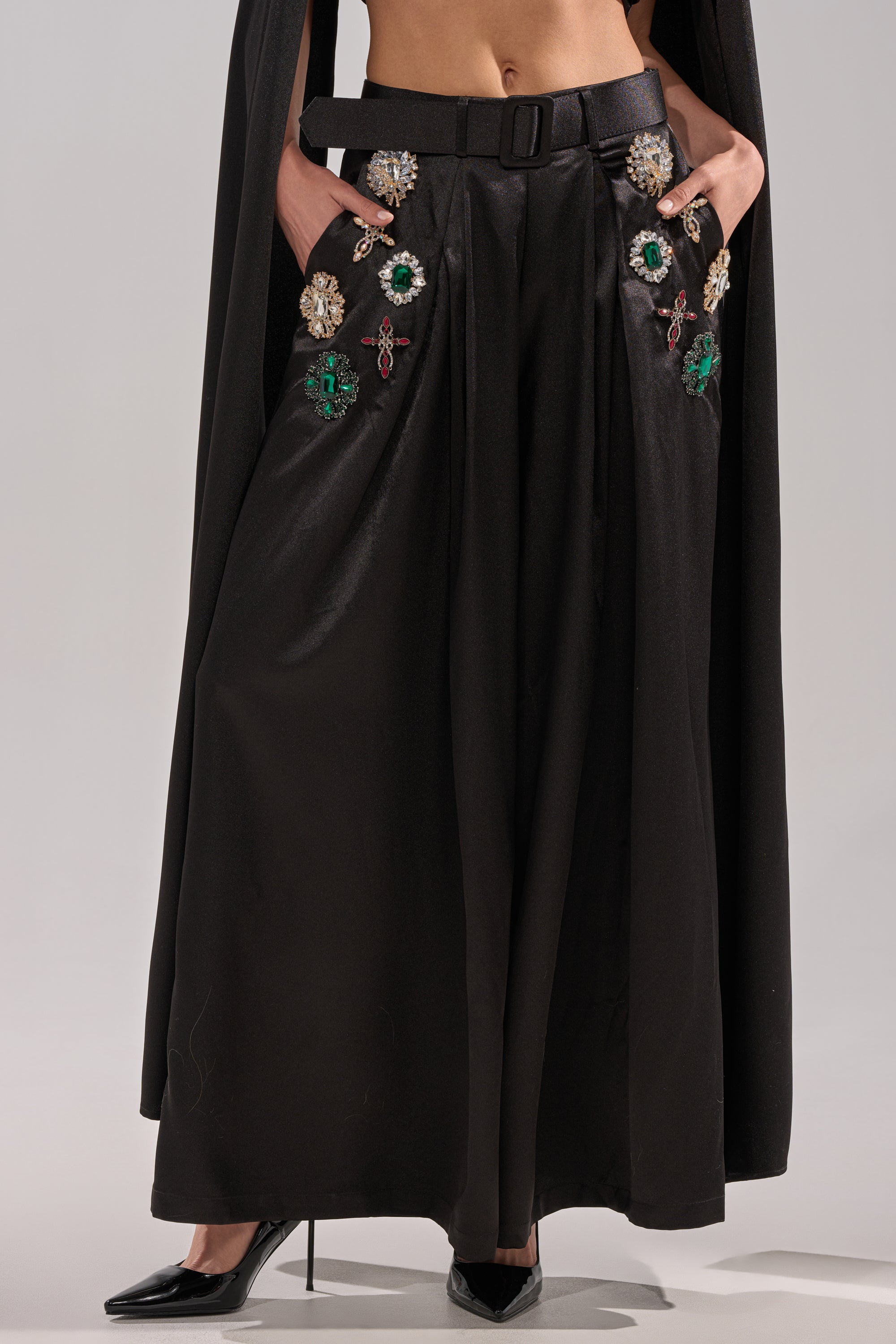 A person wears the AFTER DARK TROUSER—high-rise, wide-leg pants with jeweled embellishments by the pockets—paired with a black belt, black cape, and black pointed-toe heels. Their hands rest in their pockets.