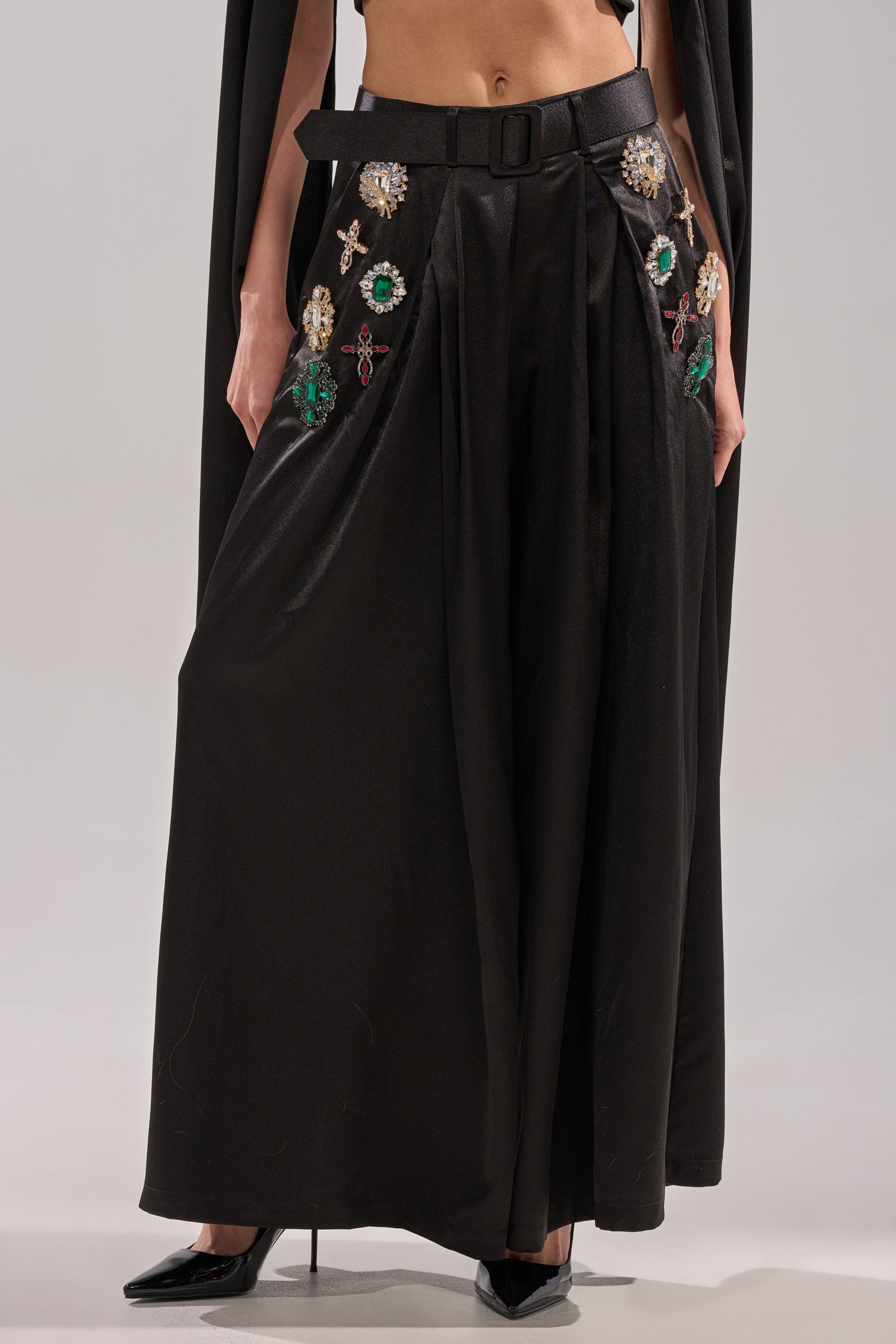 A person wears the AFTER DARK TROUSER, featuring satin wide-leg styling and rhinestone brooch details, paired with black pointed-toe heels and a sleek black top for an elegant, modern look.