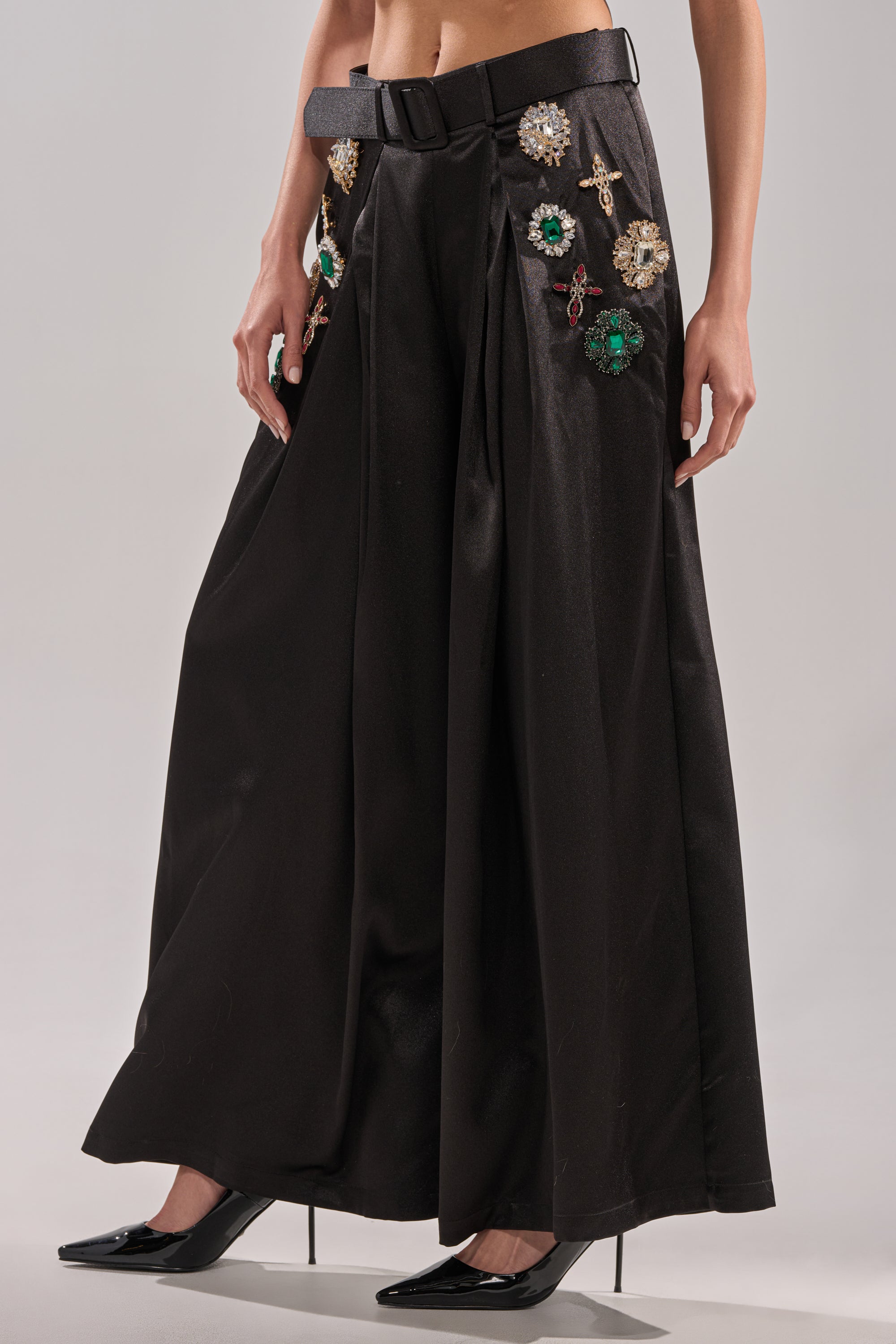 A person wears the AFTER DARK TROUSER with rhinestone brooch details, styled with a black belt and pointed heels, standing before a neutral backdrop.